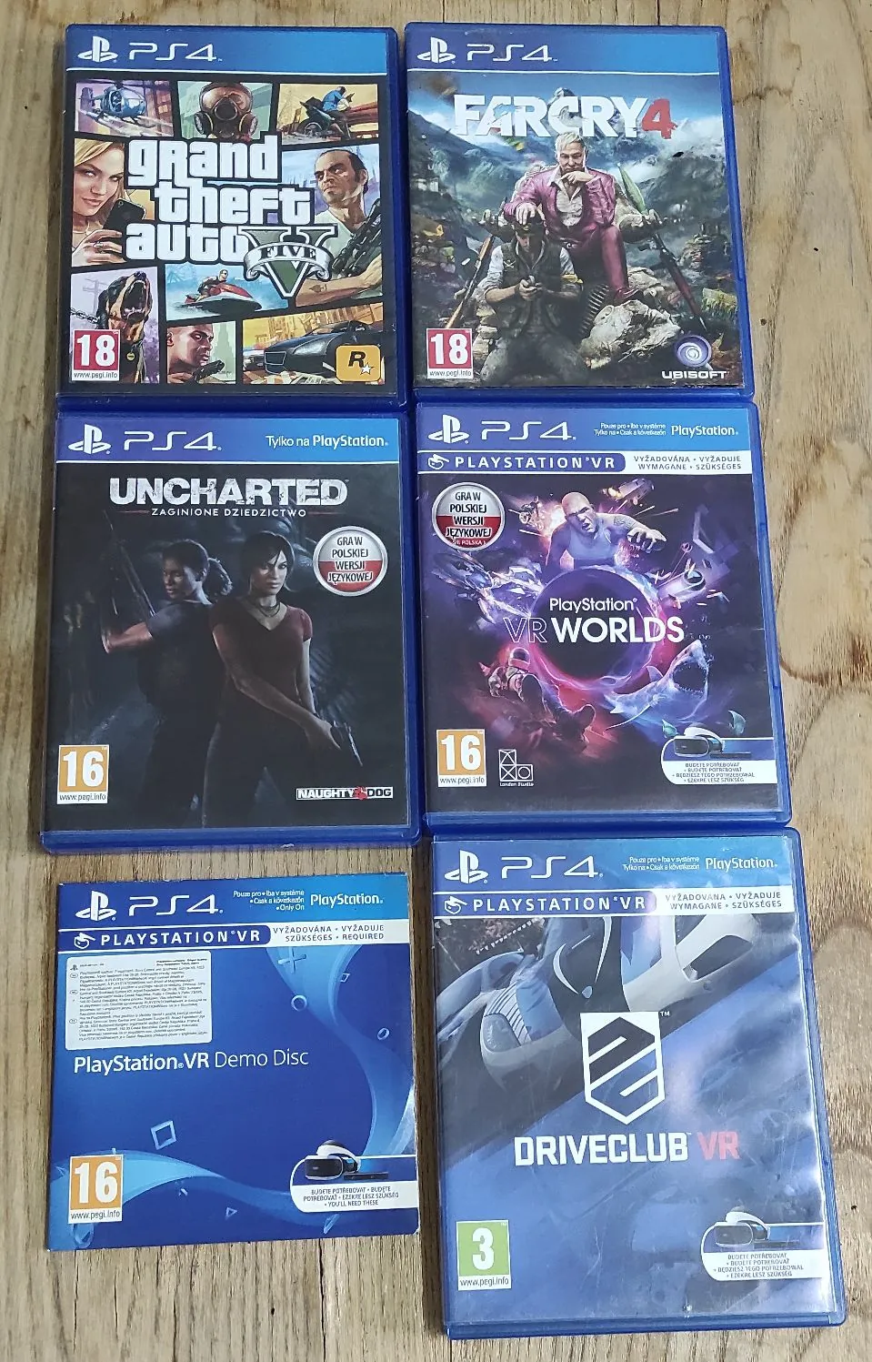 Sześć pudełek z grami na konsolę PlayStation 4 ułożonych w dwóch kolumnach po trzy sztuki na drewnianym blacie. W lewej kolumnie od góry: Grand Theft Auto V, Uncharted: Zaginione Dziedzictwo oraz PlayStation VR Demo Disc. W prawej kolumnie od góry: Far Cry 4, PlayStation VR Worlds oraz Driveclub VR. Na okładkach widoczne są logotypy PS4, klasyfikacje wiekowe PEGI oraz tytuły gier.