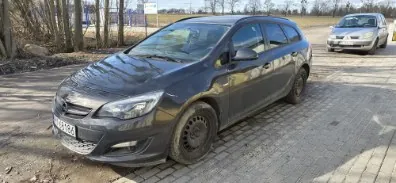 Ciemnoszary samochód osobowy typu kombi zaparkowany na brukowanej powierzchni. Widoczna tablica rejestracyjna z numerem WU 12345. W tle drugi samochód osobowy (srebrny sedan), drzewa, droga oraz tablica informacyjna. Niebo jest bezchmurne.