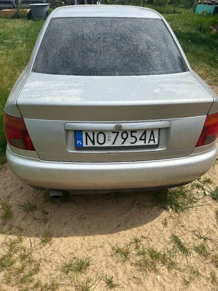 Tylny widok srebrnego samochodu osobowego zaparkowanego na piaszczystym podłożu z kępami trawy. Samochód jest zakurzony. Widoczna jest polska tablica rejestracyjna o numerze NO 7954A.