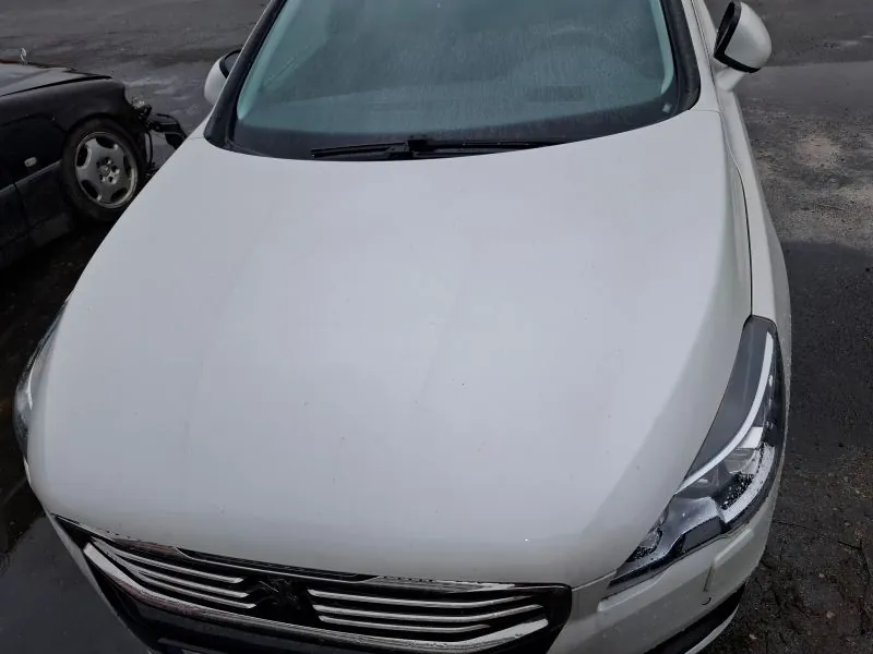 Biały samochód marki Peugeot z przodu, widoczna maska, reflektory i atrapa chłodnicy z logo. W tle po lewej stronie czarny uszkodzony samochód z widocznym przednim kołem i zdeformowaną częścią nadwozia. Tło stanowi asfaltowy parking.