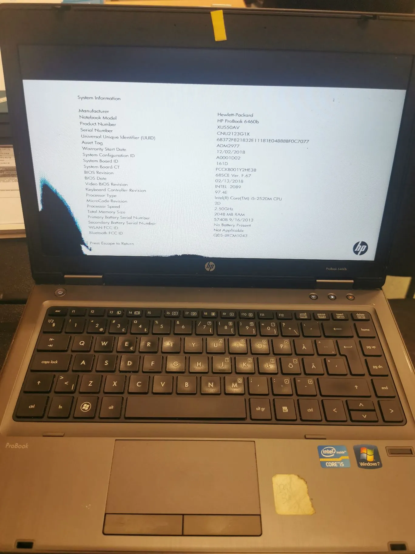 Zdjęcie otwartego laptopa marki HP ProBook 6460b. Na ekranie wyświetlone jest okno 'System Information' z danymi technicznymi urządzenia, takimi jak producent (Hewlett-Packard), model (HP Probook 6460b), numer seryjny oraz specyfikacja procesora (Intel Core i5-2520M CPU). Poniżej ekranu widoczna jest klawiatura w układzie QWERTY oraz touchpad. Na obudowie obok touchpada znajdują się naklejki 'Intel Core i5' oraz 'Windows 7'.
