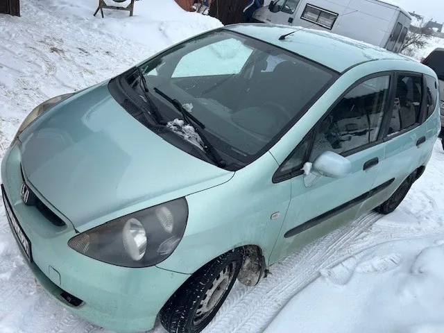 Jasnoniebieski samochód marki Honda zaparkowany na śniegu. Na przedniej szybie widoczne są resztki śniegu. W tle biały pojazd przyczepowy oraz zasneżony teren z budynkami i śladami opon.