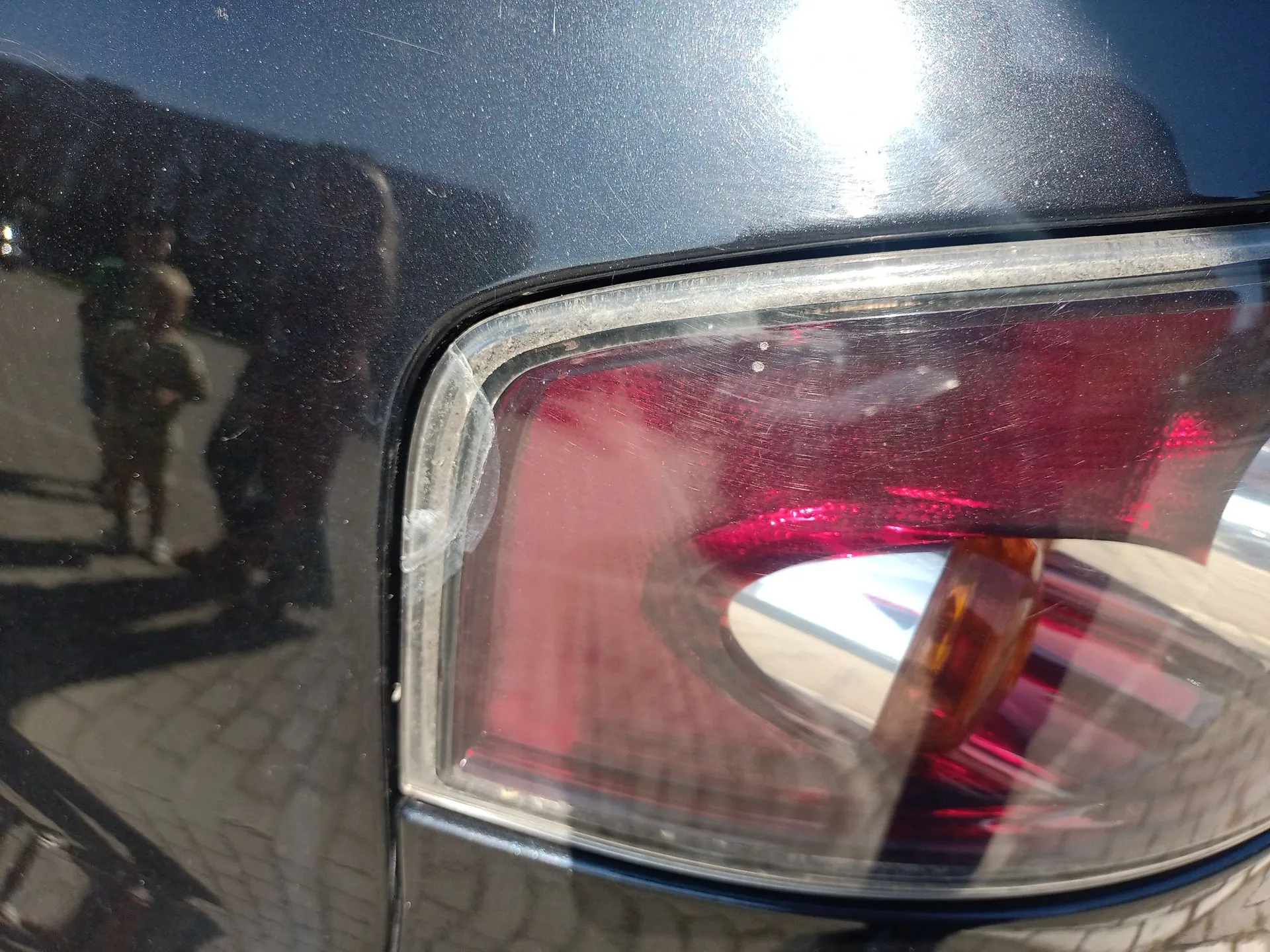 Zbliżenie na tylną lampę samochodu w kolorze czarnym. Lampa ma czerwony klosz z widocznymi zarysowaniami na powierzchni. Wewnątrz lampy widoczny jest pomarańczowy element kierunkowskazu. Na lakierze samochodu wokół lampy widoczne są drobne zarysowania oraz odbicie sylwetki osoby.
