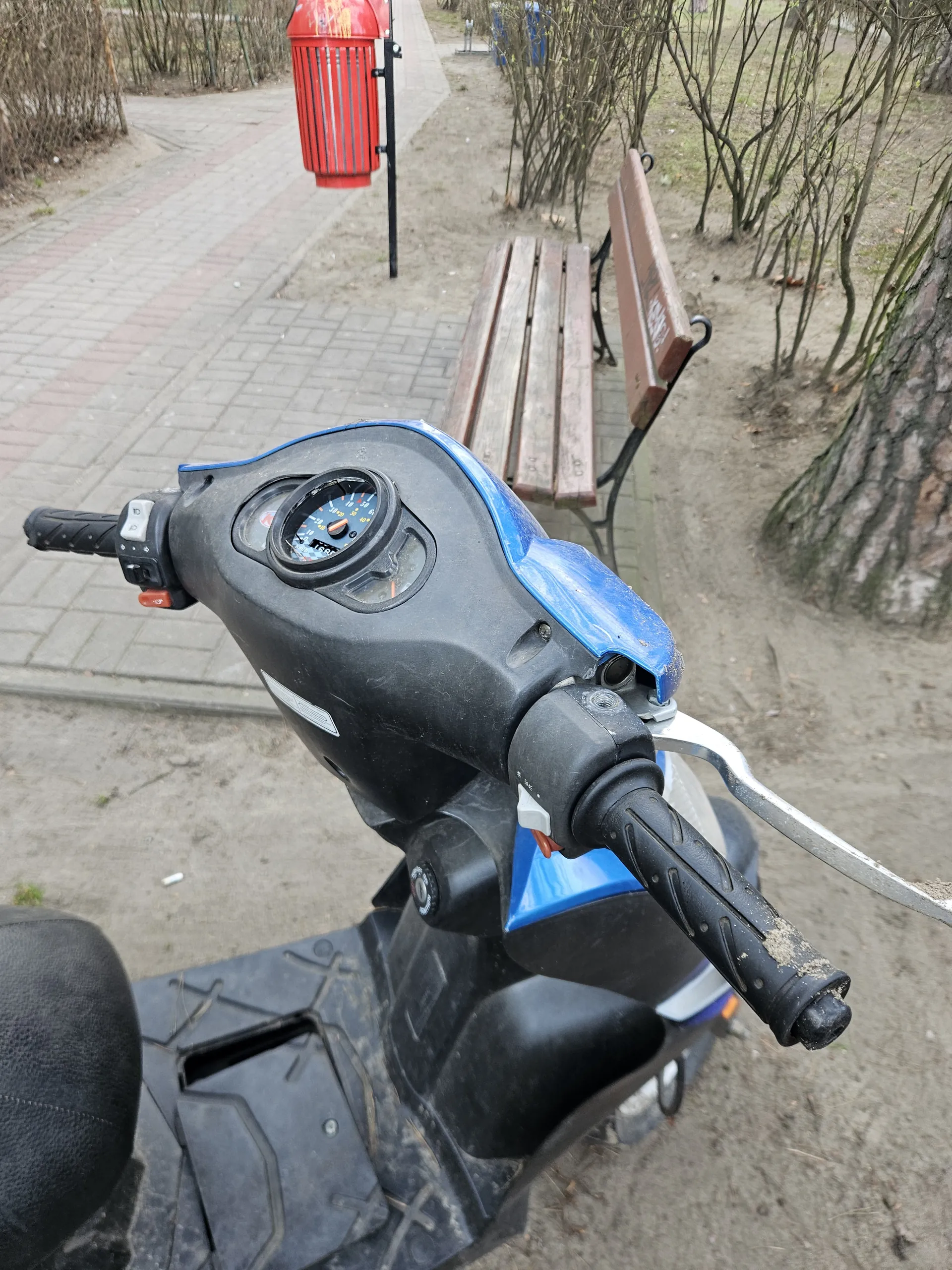 Zbliżenie na kierownicę skutera w kolorze czarno-niebieskim z licznikiem prędkości, czarnymi uchwytami pokrytymi kurzem i przyciskami sterującymi. W tle widoczna drewniana ławka z napisem 200, czerwony kosz na śmieci, nagie krzewy, pień drzewa oraz brukowana ścieżka. Całość sfotografowana z góry na tle ziemi i ścieżki.
