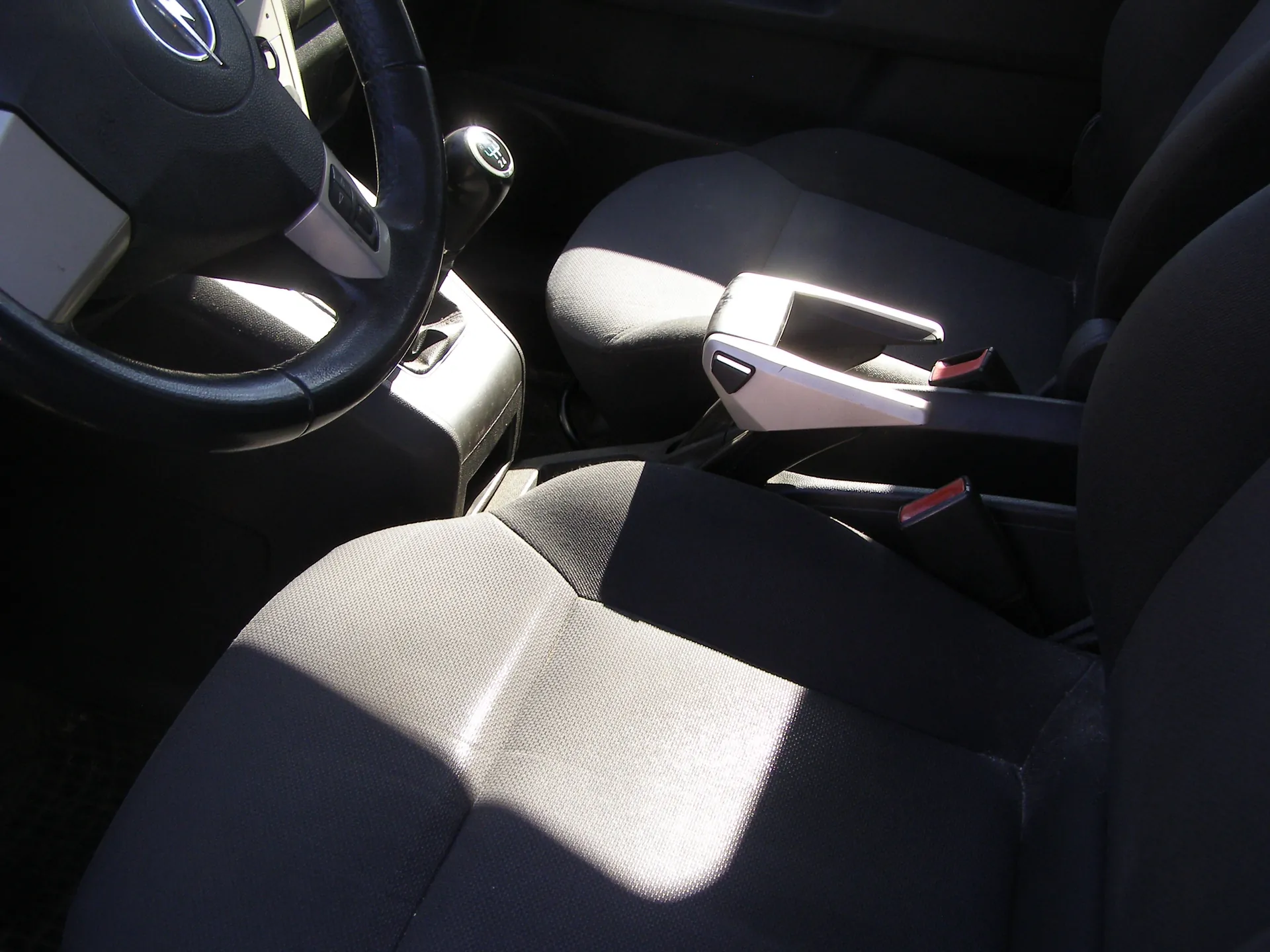 Wnętrze samochodu z widocznym kierownicą z logo po lewej stronie, dźwignią zmiany biegów, czarnymi siedzeniami z szarymi wstawkami oraz centralną konsolą z hamulcem ręcznym. Siedzenia mają pasy bezpieczeństwa z czerwonymi elementami. Światło słoneczne rzuca cienie na siedzenia.
