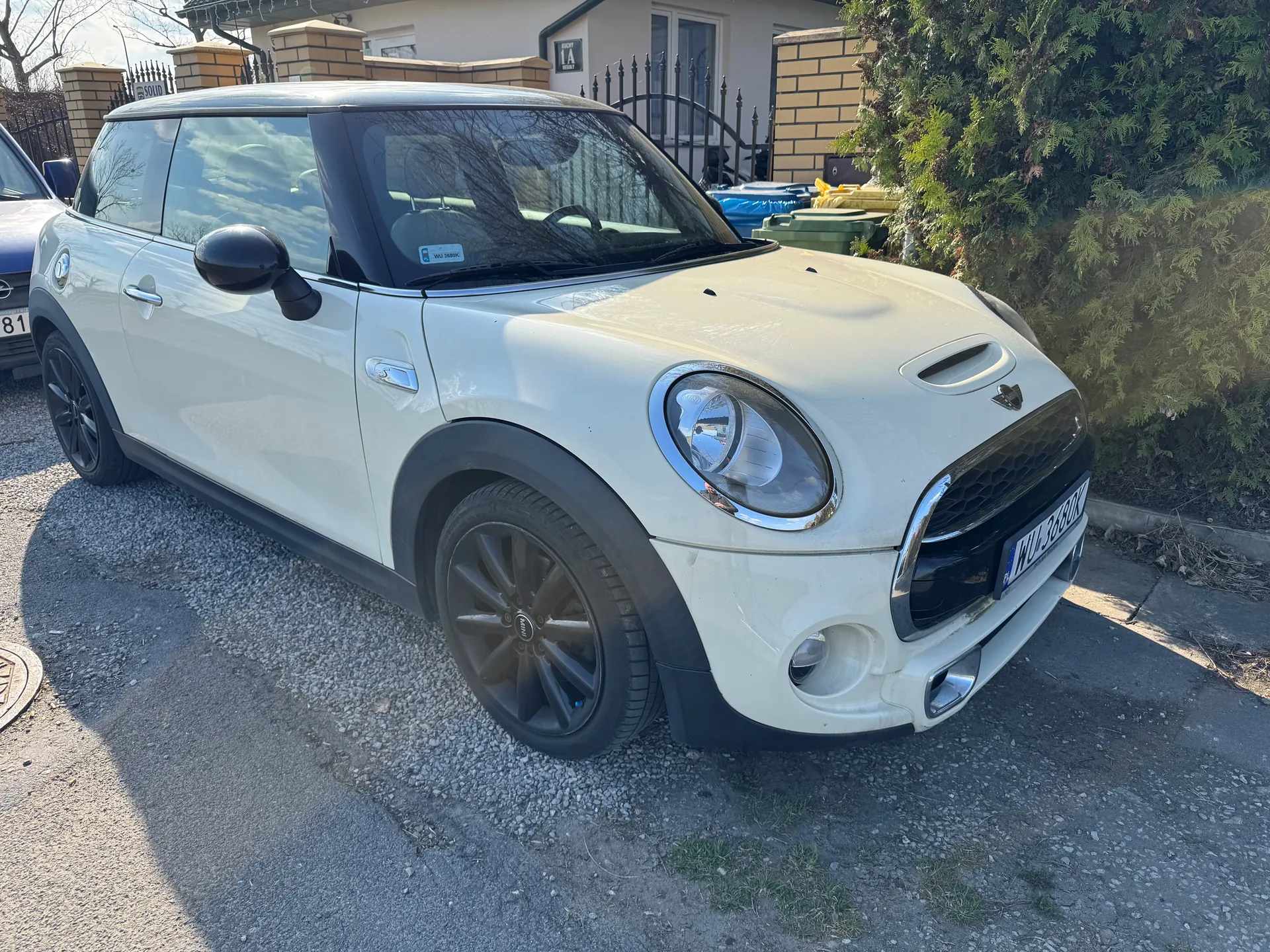 Biały samochód Mini Cooper zaparkowany na podjeździe z żwiru. Widoczne są czarne felgi, okrągłe reflektory i tablica rejestracyjna WU 3680K. W tle dom z ceglanym murem, czarnym metalowym ogrodzeniem oraz zielony krzew po prawej stronie. Częściowo widoczny inny samochód z tablicą rejestracyjną kończącą się na 81 po lewej stronie. Słoneczny dzień, cienie wskazują na jasne oświetlenie.