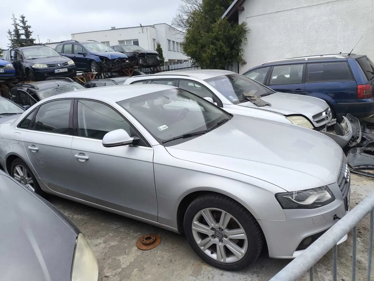 Srebrny samochód osobowy marki Audi na pierwszym planie, otoczony innymi uszkodzonymi pojazdami. W tle białe budynki, drzewa i pochmurne niebo. Widoczna tablica rejestracyjna na ciemnym samochodzie w tle: KMA 3107. Na ziemi leży rdzewiejący dysk hamulcowy, a na pierwszym planie metalowe ogrodzenie. Samochody są zaparkowane na betonowej powierzchni.