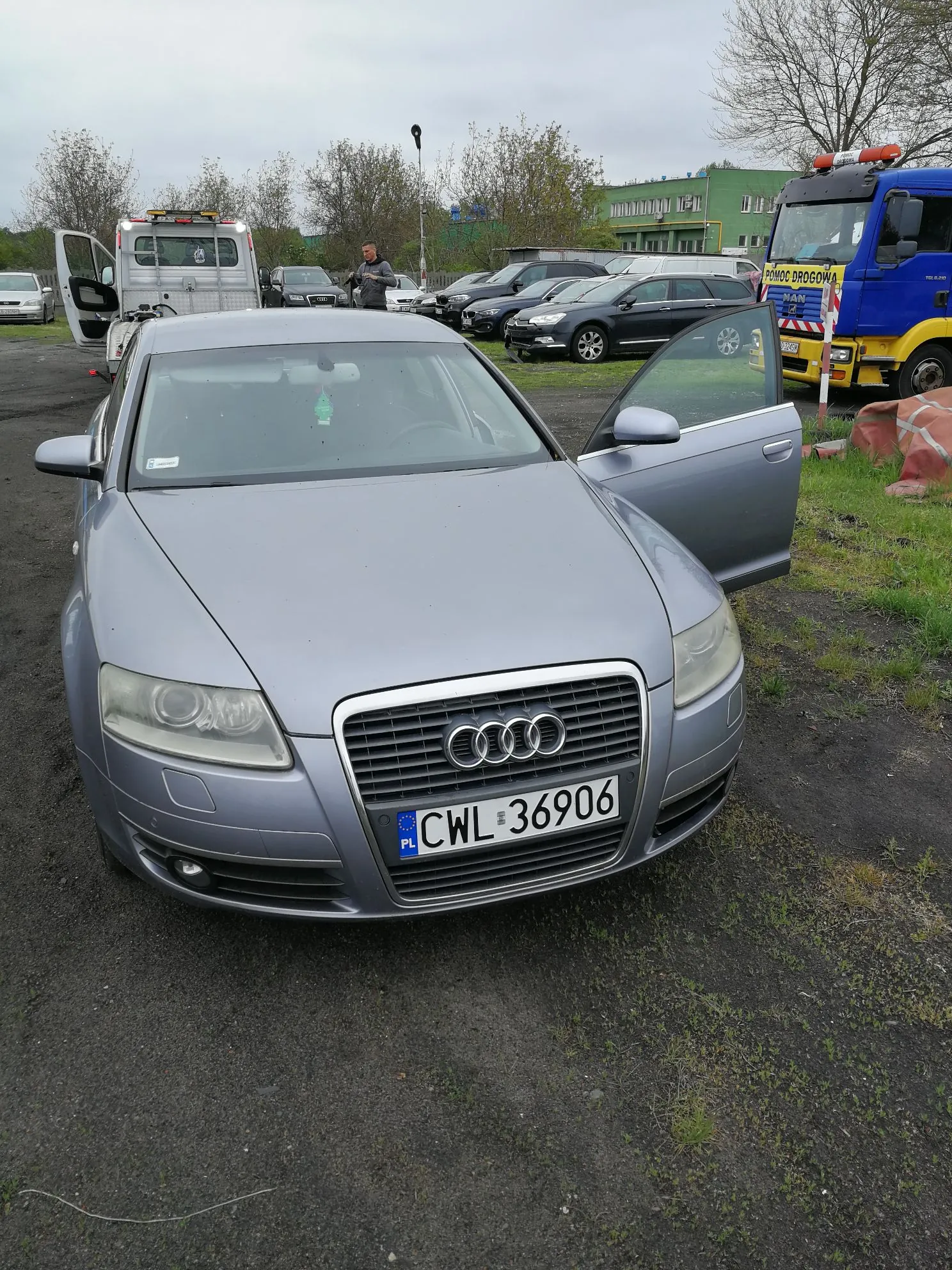 Srebrny samochód marki Audi zaparkowany przodem do widza na żwirowym placu. Pojazd ma otwarte drzwi kierowcy. Na przodzie widoczna jest polska tablica rejestracyjna o numerze CWL 36906. W tle znajdują się inne zaparkowane samochody, w tym żółty samochód pomocy drogowej z napisem 'POMOC DROGOWA' oraz zielony budynek.