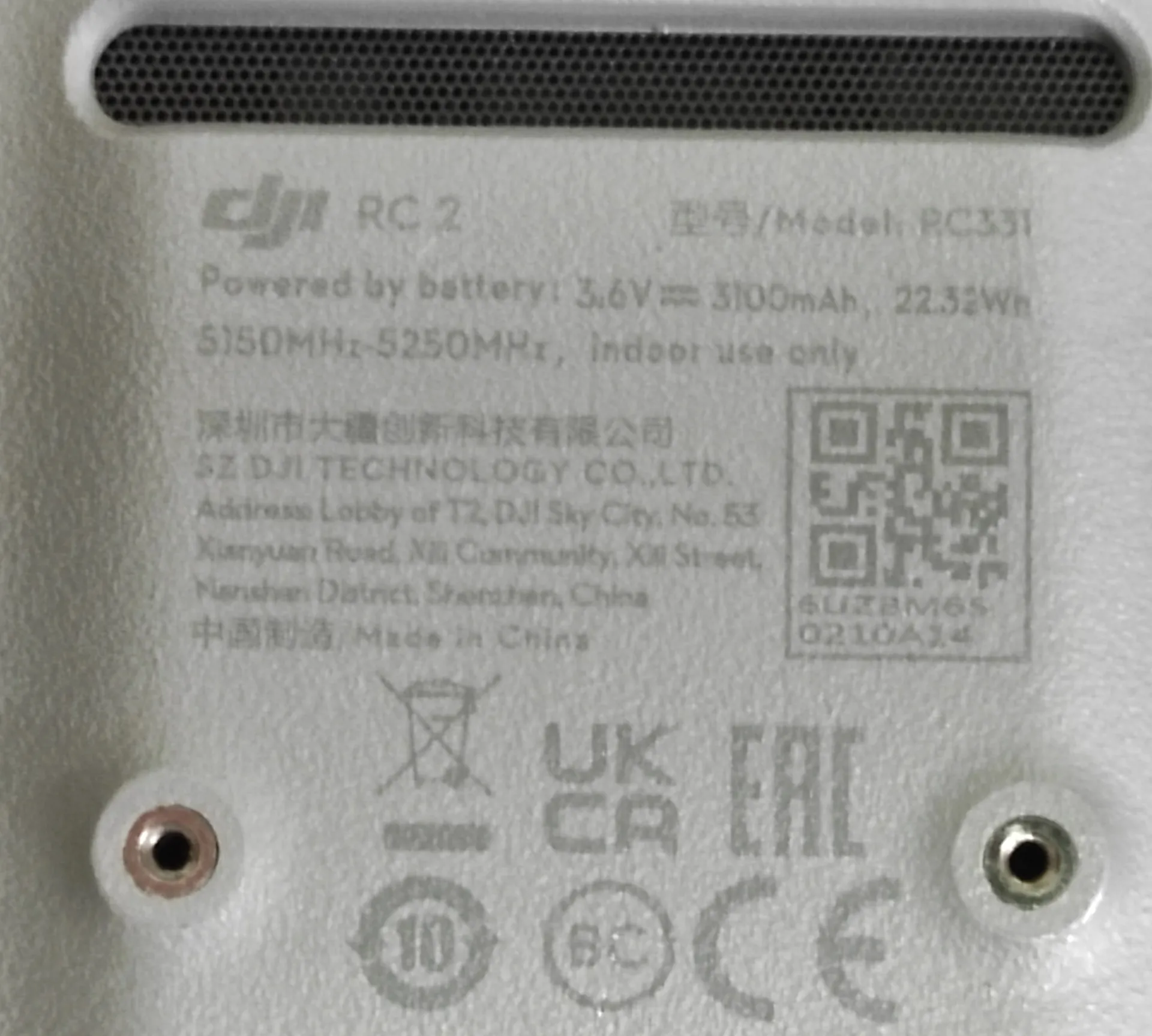 Zbliżenie na tylną obudowę urządzenia DJI RC 2. Widoczny jest tekst: 'DJI RC 2', 'Model: RC331', 'Powered by battery: 3.6V 3100mAh, 22.32Wh', '5150MHz-5250MHz, indoor use only'. Poniżej znajduje się nazwa producenta 'SZ DJI TECHNOLOGY CO., LTD.' wraz z adresem w Shenzhen, Chiny oraz informacja 'Made in China'. Po prawej stronie widnieje kod QR i numer seryjny. Na dole znajdują się symbole certyfikacji: przekreślony kosz, UKCA, EAC, CE oraz inne znaki techniczne. W górnej części widoczny jest fragment głośnika z siatką, a po bokach dwa otwory montażowe z metalowymi wkładami.