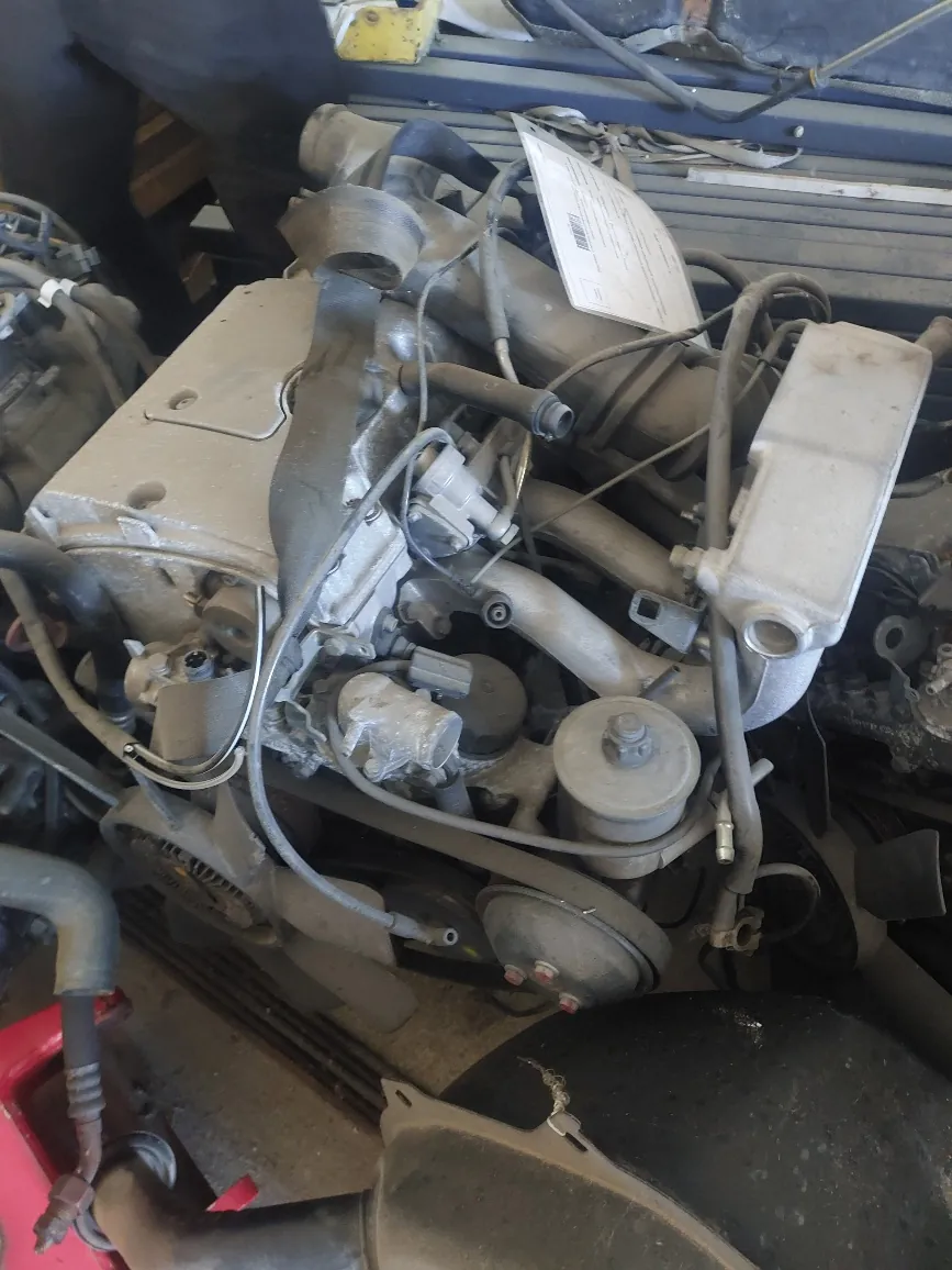 Zbliżenie na zakurzony, używany silnik spalinowy z zamontowanymi przewodami, rurami i elementami mechanicznymi, leżący na innym podzespole samochodowym.