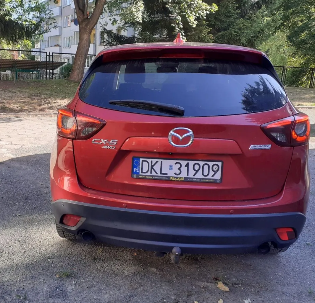 Tylny widok czerwonego samochodu marki Mazda model CX-5 AWD. Pojazd posiada polską tablicę rejestracyjną o numerze DKL 31909. Na klapie bagażnika widoczne są emblematy 'CX-5 AWD', logo Mazdy oraz napis 'SKYACTIV'. Samochód zaparkowany jest na szutrowym podłożu, w tle widoczne są drzewa oraz fragment białego budynku mieszkalnego.
