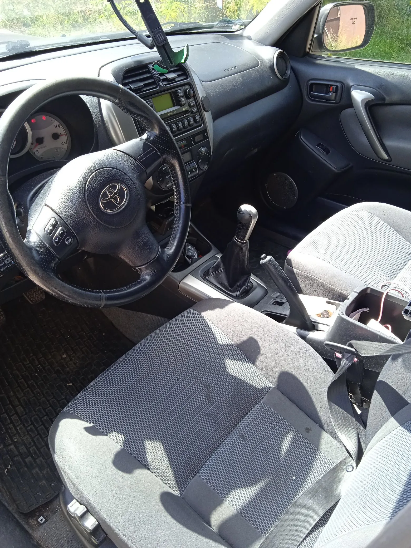 Wnętrze samochodu z perspektywy kierowcy. Widoczna jest czarna kierownica z logo Toyoty, deska rozdzielcza z radiem i panelami sterowania, dźwignia zmiany biegów oraz dwa szare, materiałowe fotele. Na podłodze znajduje się gumowa mata. Przez przednią szybę i lusterko boczne widoczny jest fragment zieleni.