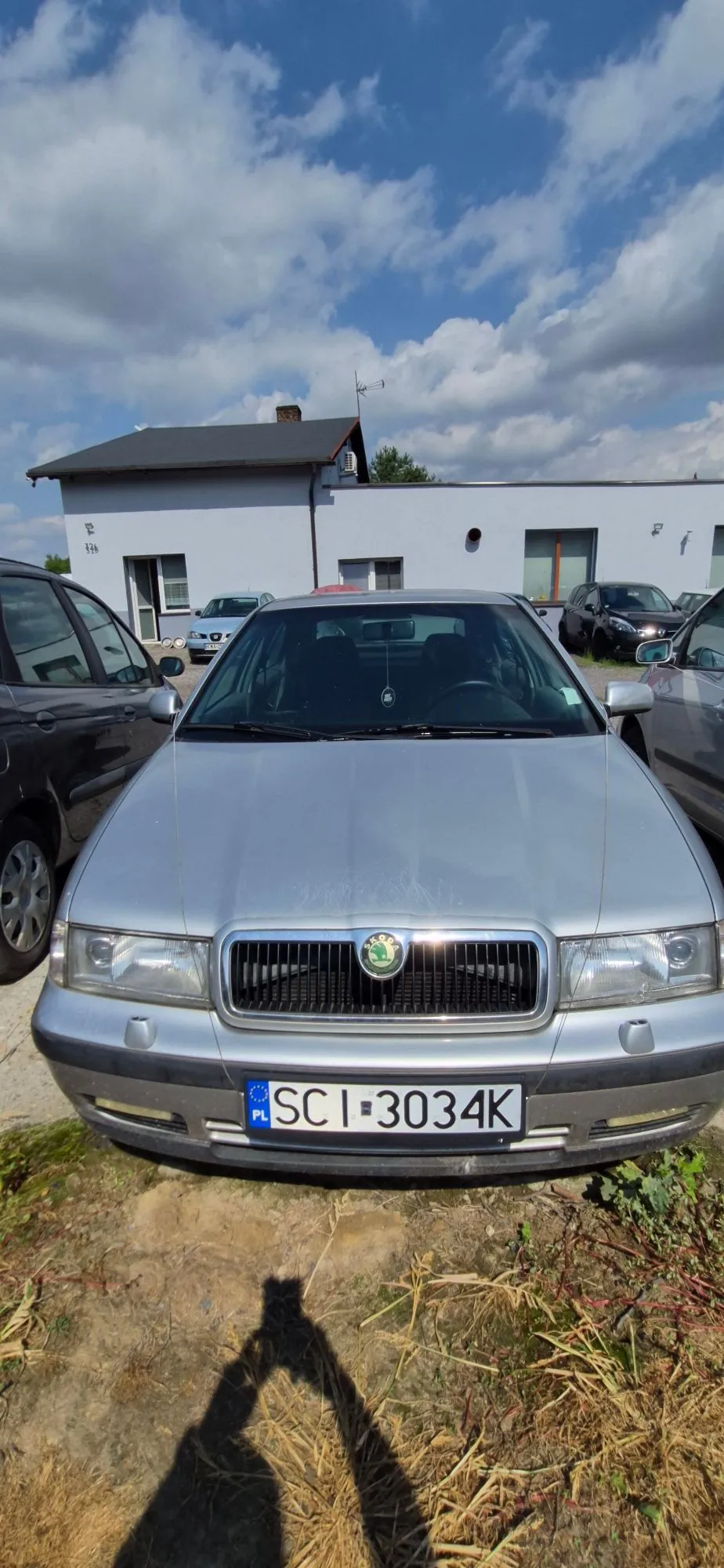 Srebrny samochód marki Skoda zaparkowany przodem do obiektywu na piaszczystym podłożu. Pojazd posiada polską tablicę rejestracyjną o numerze SCI 3034K. W tle widoczne są inne zaparkowane samochody oraz jasny, parterowy budynek pod niebem z białymi chmurami.