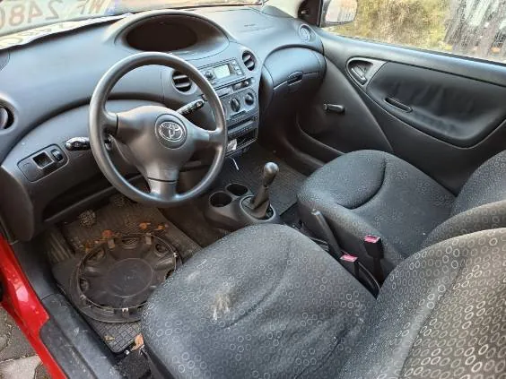 Wnętrze samochodu marki Toyota z widocznym kierownicą z logo, deska rozdzielcza z radioodtwarzaczem i przyciskami, dźwignią zmiany biegów, czarnymi siedzeniami z wzorem okręgów oraz uszkodzonym dywanikiem pod kierowcą. Drzwi pasażerskie są otwarte, widoczna czerwona karoseria. W tle za oknem drzewa i fragment budynku.