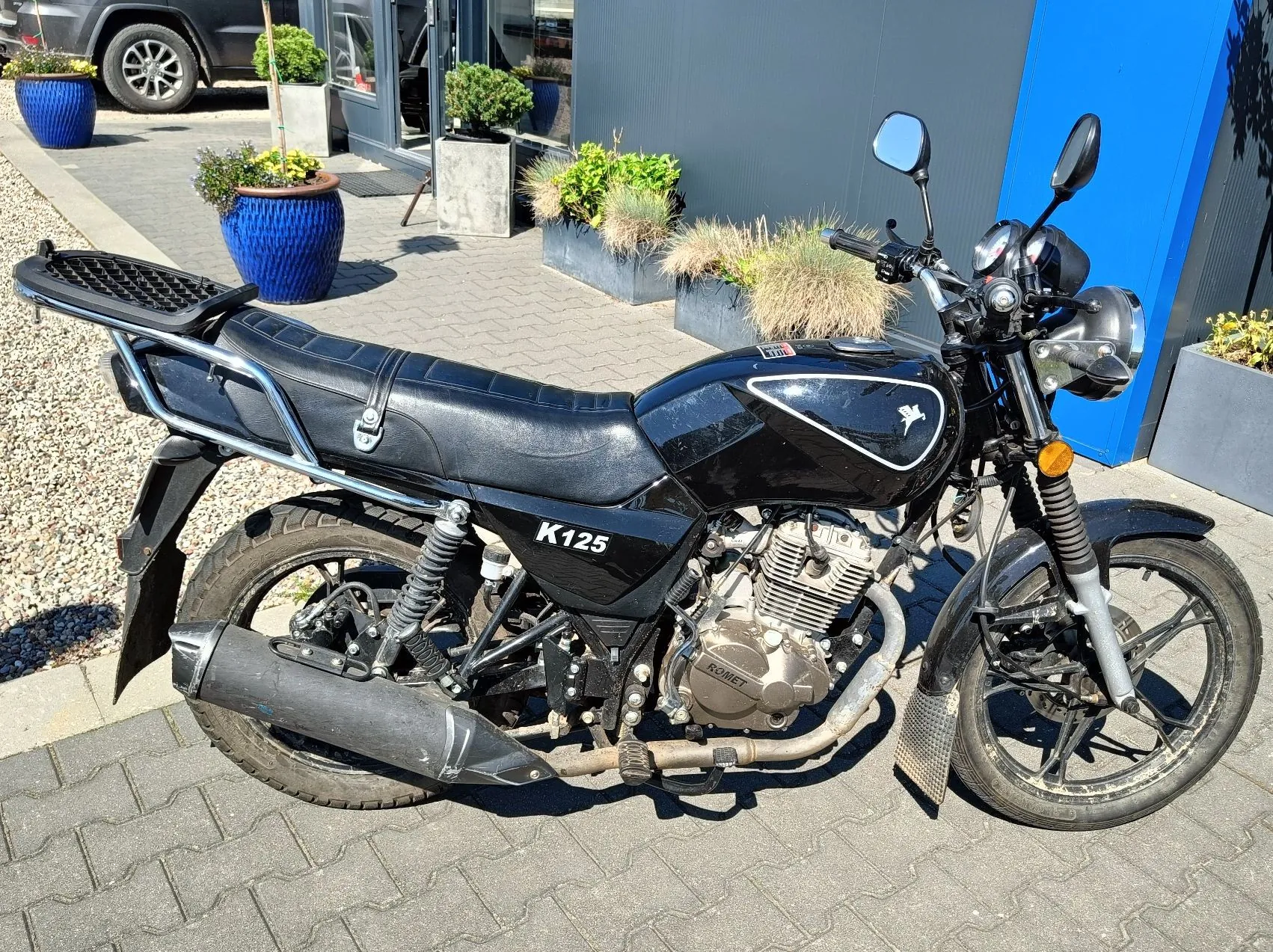 Czarny motocykl model K125 zaparkowany bokiem na kostce brukowej przed budynkiem. Pojazd posiada czarną kanapę, metalowy bagażnik z tyłu, srebrny silnik z napisem KOMET oraz czarną rurę wydechową. W tle widoczne są niebieskie donice z roślinami oraz fragment niebieskiej ściany budynku.
