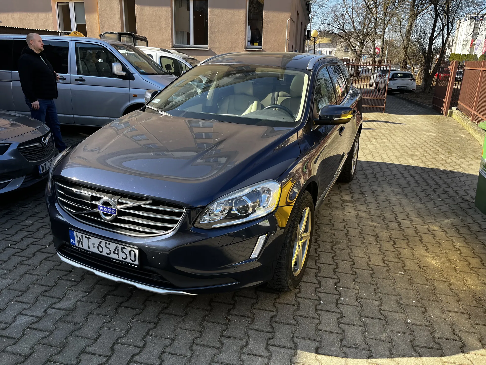 Ciemnoniebieski samochód Volvo XC60 z tablicą rejestracyjną WT 65450 (PL) zaparkowany na brukowanym placu. W tle srebrny van, osoba w czarnej kurtce i niebieskich spodniach, budynki o beżowej elewacji, metalowe ogrodzenie oraz drzewa bez liści. Widoczne są również inne zaparkowane pojazdy i jasne niebo.