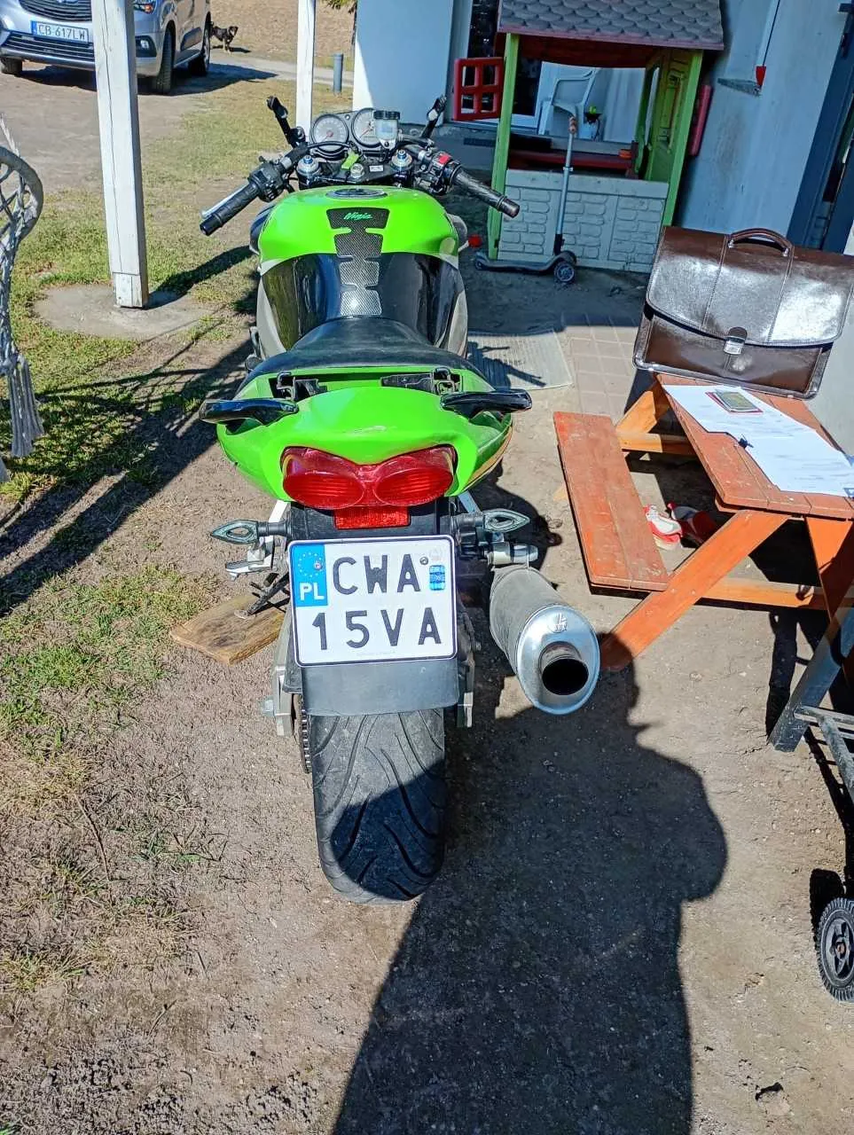 Zdjęcie przedstawia z tyłu zielony motocykl z tablicą rejestracyjną PL CWA 15VA. W tle widoczny jest drewniany stolik piknikowy z brązową teczką, dokumentami i telefonem komórkowym. Po prawej stronie znajduje się mały domek zabaw z zielonymi drzwiami, a w oddali srebrny samochód z tablicą CB 617L. Tło stanowi trawnik i podwórze z betonowymi elementami.