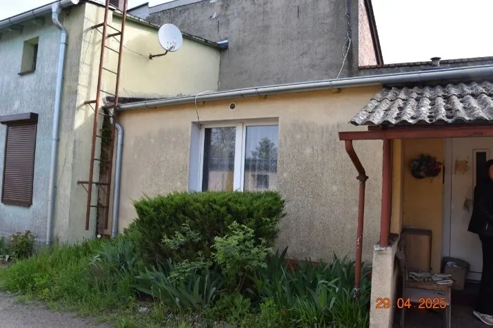 Zdjęcie fragmentu budynku mieszkalnego z jasną, tynkowaną elewacją. Widoczna jest metalowa drabina przymocowana do ściany, antena satelitarna na dachu oraz okno z białymi ramami. Przed budynkiem rośnie gęsty, zielony krzew i roślinność trawiasta. Po prawej stronie znajduje się zadaszone wejście z brązowymi wspornikami. W prawym dolnym rogu widnieje czerwona data: 29.04.2025.