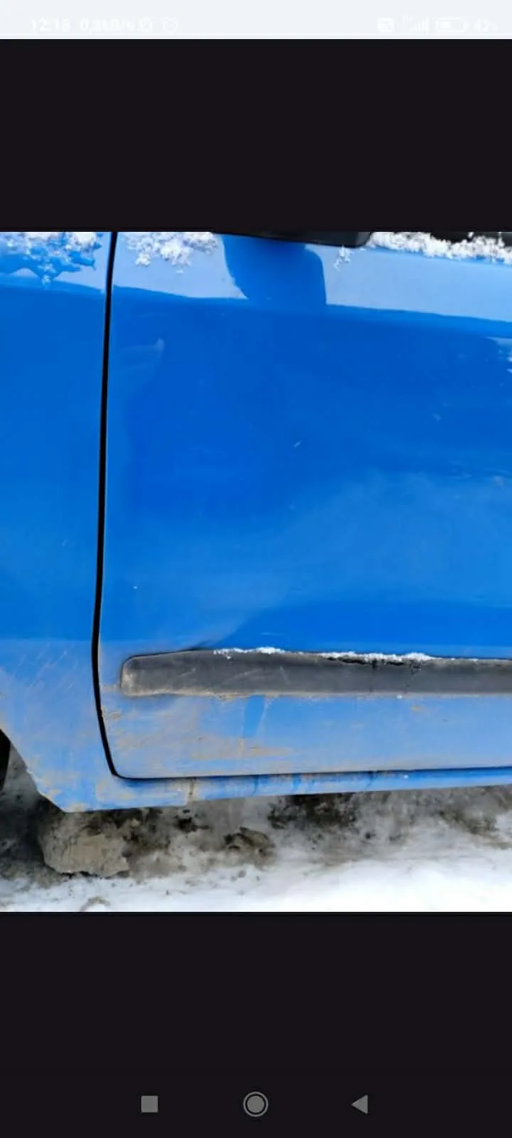 Boczna część niebieskiego samochodu z widocznym czarnym zderzakiem, śniegiem na powierzchni i błotem na dolnej części. Tło z zasnieżonym terenem.