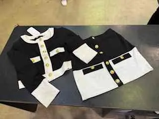 Dwa czarno-białe stroje leżące na ciemnym stole. Każdy strój składa się z czarnej kurtki z białym kołnierzem i białymi elementami, złotymi guzikami oraz białych spodni lub spódnicy z czarnym paskiem i złotymi guzikami. Tło stanowi betonowa podłoga, w prawym górnym rogu widoczna jest część nogi osoby.