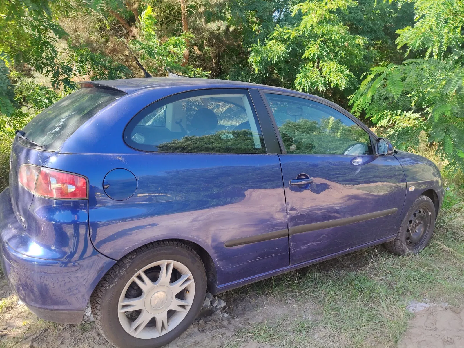 Niebieski samochód osobowy typu hatchback zaparkowany na piaszczystym podłożżu na tle gęstych, zielonych krzewów i drzew. Widoczny jest profil samochodu od strony pasażera, z widocznymi wgnieceniami na drzwiach i tylnym błotniku.