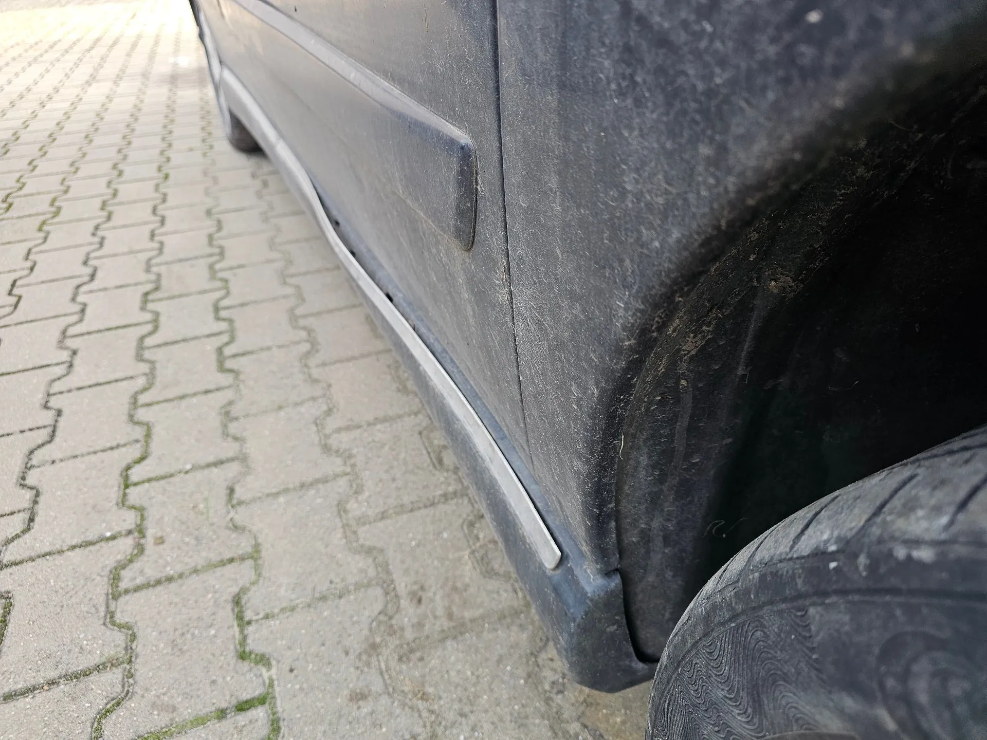 Zbliżenie na dolną część boku ciemnego samochodu, zaparkowanego na kostce brukowej. Widoczny jest próg auta z metalowym elementem wykończeniowym oraz fragment tylnego nadkola i opony.