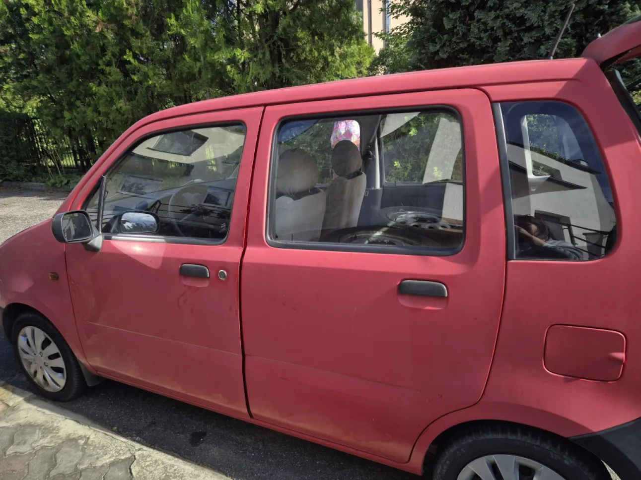 Boczny widok czerwonego, pięciodrzwiowego samochodu osobowego zaparkowanego na podjeździe. Wewnątrz pojazdu widoczne są jasne zagłówki foteli. W tle znajdują się zielone drzewa i krzewy.