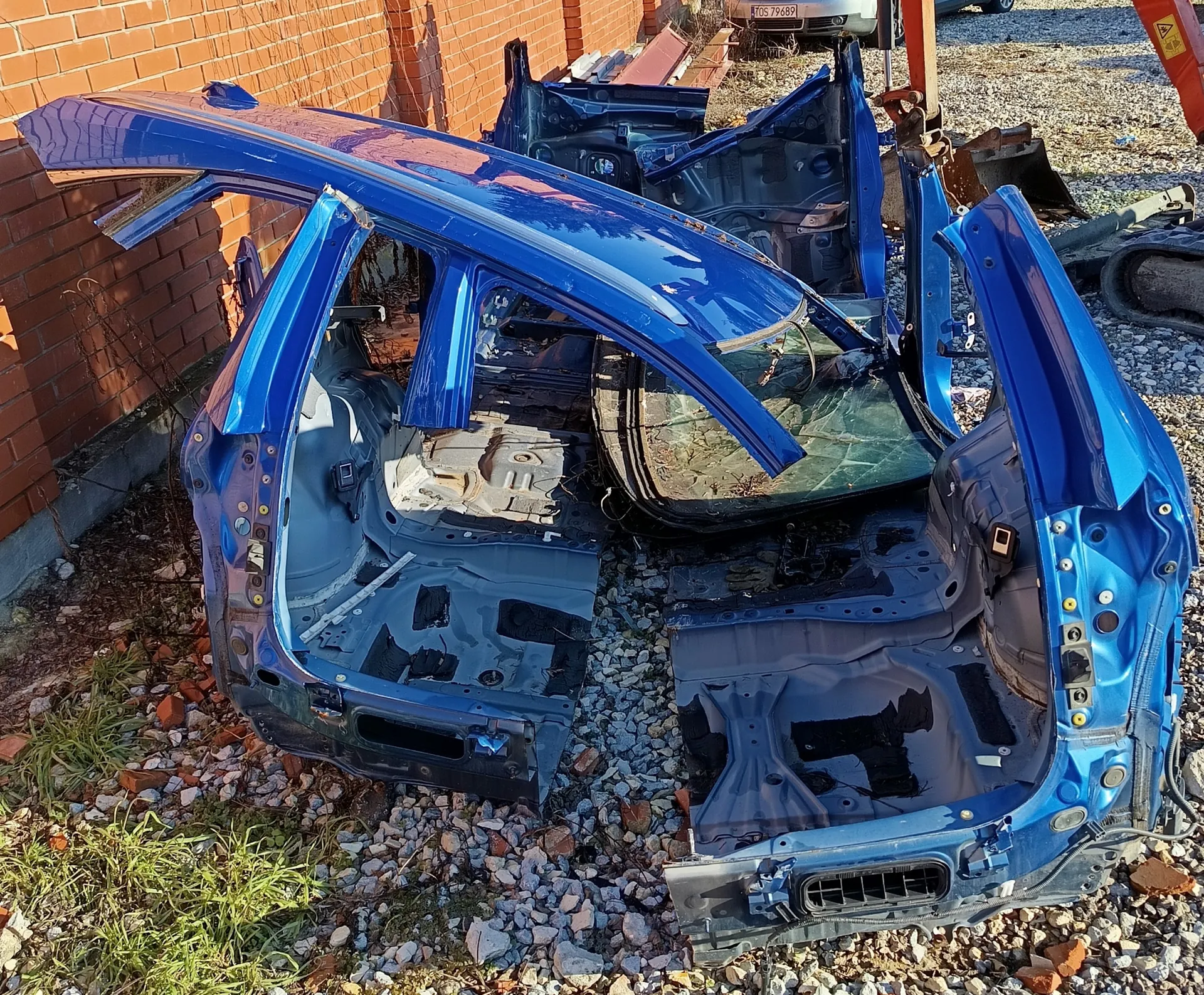 Niebieski wrak samochodu bez drzwi i wnętrza, z uszkodzonym przednim szybą, położony na żwirowym podłożu. W tle ceglana ściana, fragment innej pojazdu z widoczną tablicą rejestracyjną TOS 79689 oraz części maszyny budowlanej. Na ziemi rosną niewielkie łaty trawy, a wokół rozrzucone są kawałki cegieł.