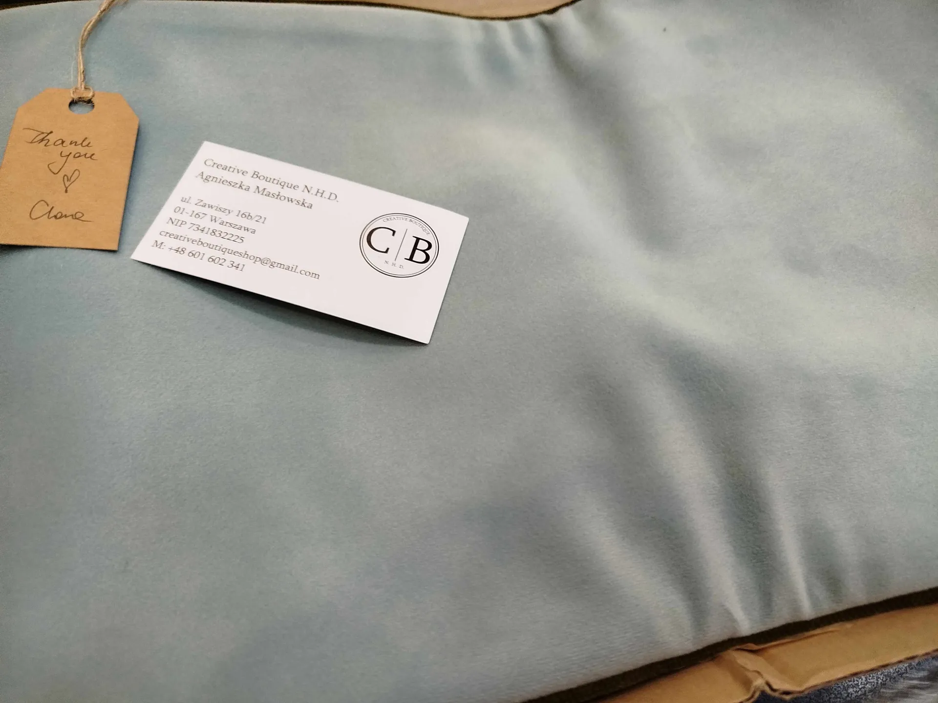 Zbliżenie na jasnoniebieską tkaninę z brązową etykietą zawieszoną na sznurku. Na etykiecie ręcznie napisano 'Thank you' oraz serce, poniżej imię 'Clare'. Obok znajduje się biała wizytówka z napisem 'Creative Boutique N.H.D. Agnieszka Masłowska', adresem 'ul. Zawiszy 16b/21 01-167 Warszawa', numerem NIP '7341832225', adresem e-mail 'creativeboutiqueshop@gmail.com' oraz numerem telefonu 'M: +48 601 602 341'. W prawym górnym rogu wizytówki umieszczono okrągły logo z literami 'C B'.