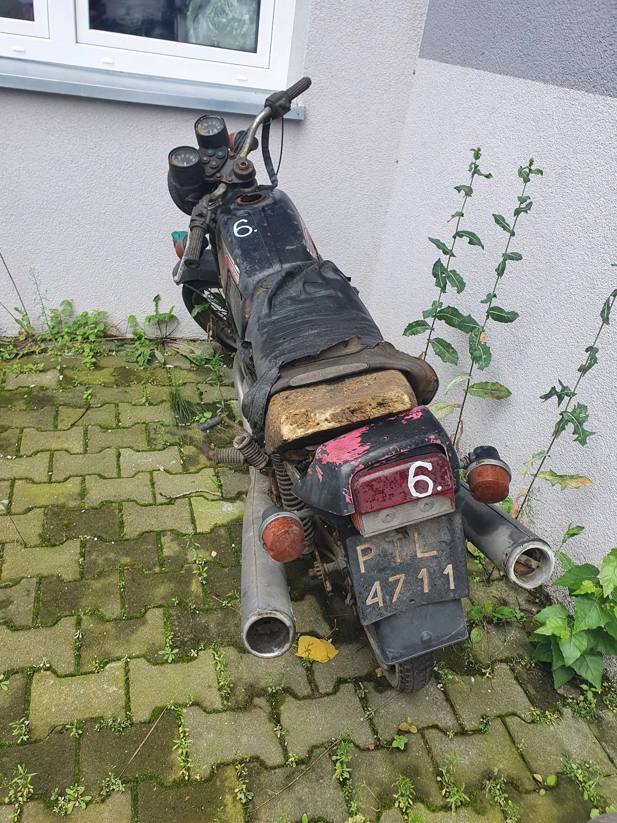 Stary, zniszczony motocykl zaparkowany na kostce brukowej porośniętej mchem. Pojazd jest czarny, z widocznymi śladami rdzy i zużycia. Na baku oraz na tylnym błotniku widnieje biała cyfra „6.”. Siedzisko jest częściowo przykryte czarną tkaniną, a pod nią znajduje się kawałek gąbki. Z tyłu znajduje się tablica rejestracyjna z napisem „PTL 4711”. Motocykl stoi na tle jasnej ściany budynku i kilku zielonych roślin.