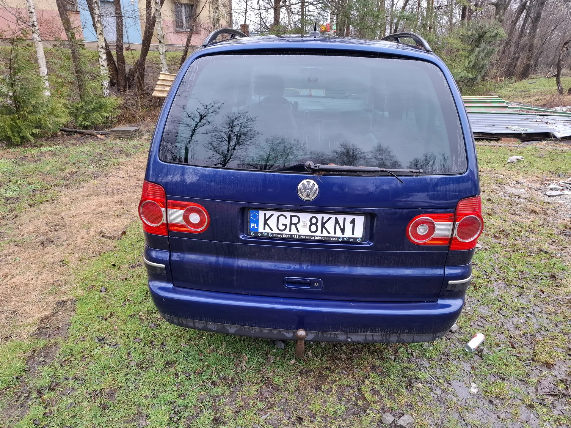 Tył niebieskiego samochodu marki Volkswagen z tablicą rejestracyjną KGR 8KN1 (PL). Samochód stoi na trawie z suchymi plamami, w tle widoczne są drzewa, krzewy oraz budynek z niebiesko-różowymi ścianami. Na ziemi obok pojazdu leży pusta puszka. Tablica rejestracyjna zawiera dodatkowy napis: Nowy Sącz 725. rocznica lokacji miasta.