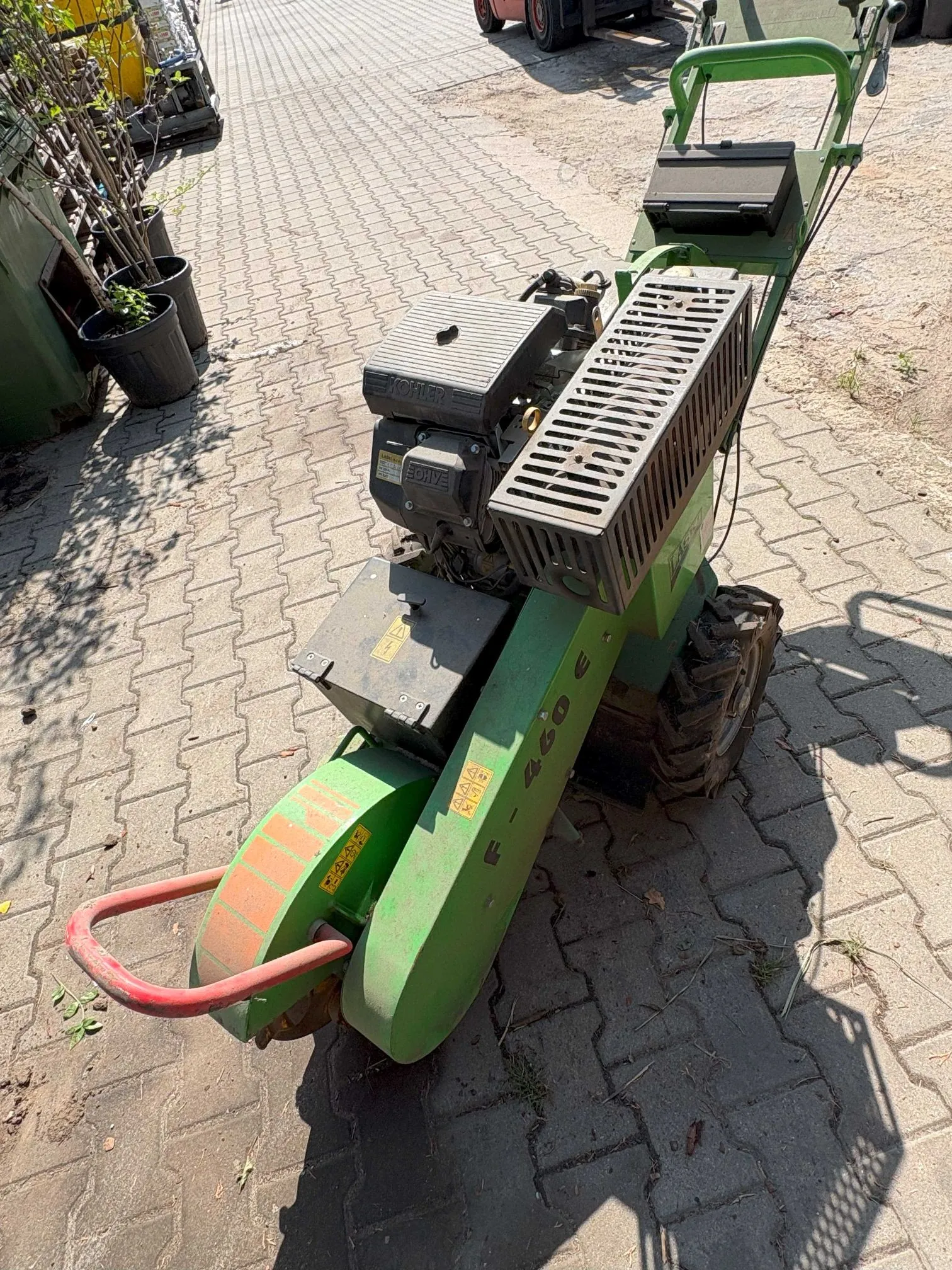 Zielona kosiarka spalinowa do pielenia lub glebogryzarka stojąca na kostce brukowej. Maszyna posiada silnik marki Kohler, metalową obudowę w kolorze zielonym z oznaczeniem 'F 460 E' oraz czerwony metalowy uchwyt z przodu. W tle widoczne są doniczki z roślinami oraz fragmenty innych urządzeń.