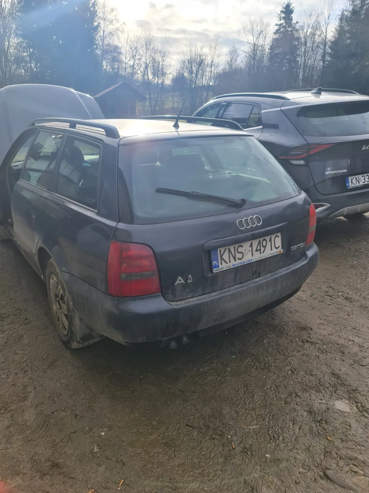Zdjęcie przedstawia ciemnoszary samochód Audi A4 kombi z otwartą klapą bagażnika i otwartą przednią klapą. Na tablicy rejestracyjnej widoczny jest numer KNS 1491C z oznaczeniem PL. Samochód jest zabrudzony błotem, stoi na błotnistym terenie. W tle znajduje się szary SUV z tablicą KN-33 oraz drzewa bez liści, niewielka budowla i częściowo zachmurzone niebo. Na tylnym zderzaku Audi widoczne są napisy A4 i 1.9 TDI.