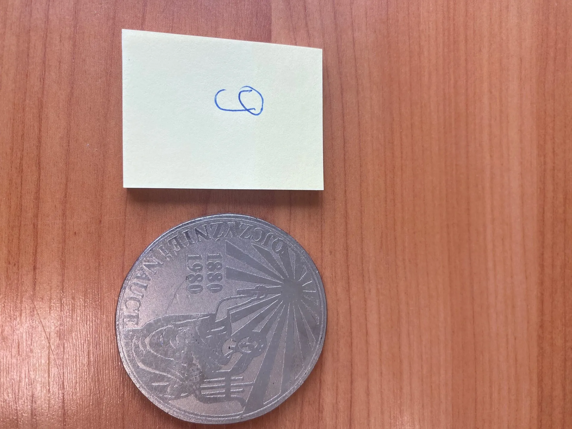 Na drewnianym tle znajduje się jasnozielony karteczkowy notatnik z niebieskim napisem "CQ" oraz duża srebrna moneta. Na monecie widoczne są liczby "1880" i "1980", napisy wokół obwodu oraz grafika przedstawiająca postać z promieniującym tłem.