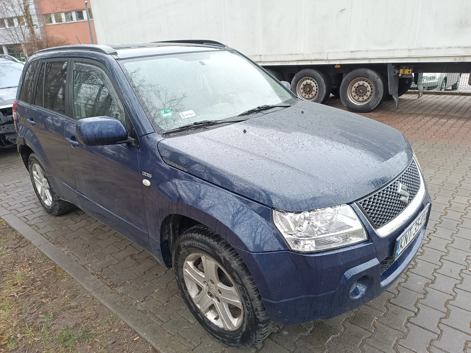 Niebieski SUV marki Suzuki z tablicą rejestracyjną KWT 3544 zaparkowany na brukowanej powierzchni. Samochód pokryty jest kroplami wody, na przedniej szybie widoczne są dwa naklejki (zielona i biała). W tle biały naczepowy ciężarówka z kołami oraz budynki z oknami. Po lewej stronie fragment innego uszkodzonego pojazdu. Na boku SUV widoczny napis DDIS.