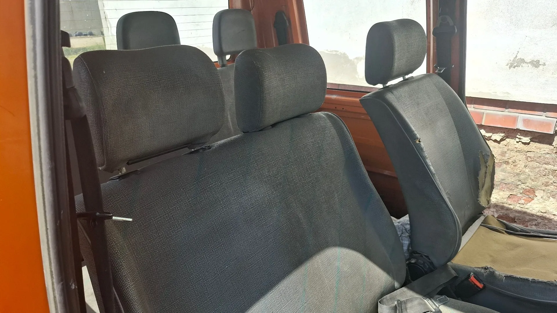 Wnętrze pomarańczowego pojazdu z rzędem szarych, materiałowych siedzeń. Na pierwszym planie widoczna jest kanapa z dwoma zagłówkami, a obok niej pojedynczy fotel z uszkodzoną, przetartą tapicerką na boku. W tle widać kolejne zagłówki siedzeń oraz fragment białej ściany budynku za oknem.