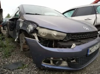 Zniszczony szary samochód marki Volkswagen z uszkodzonym przodem, w tym zdeformowaną maską, zniszczonym zderzakiem i uszkodzonym reflektorem. Widoczna tablica rejestracyjna z numerem W0729H. W tle inny uszkodzony pojazd, prawdopodobnie SUV. Scena rozgrywa się na podłożu z żwiru i trawy, z chmurnym niebem.