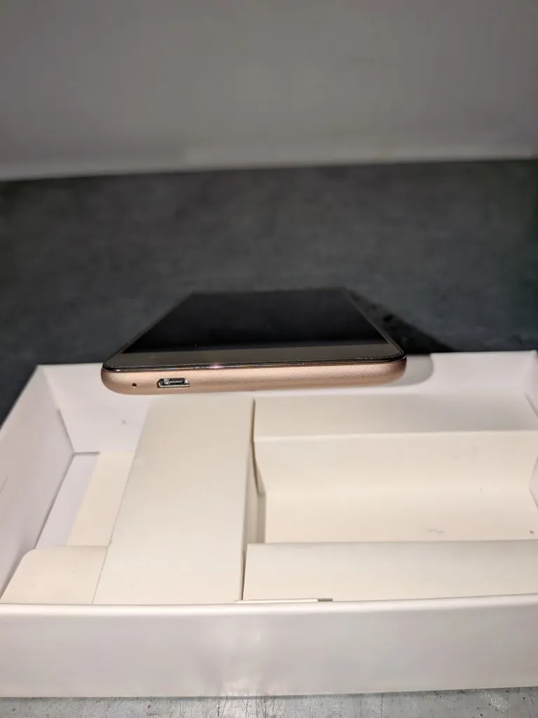 Zdjęcie przedstawia smartfon w kolorze złotym, leżący poziomo na białym, otwartym pudełku. Widoczna jest dolna krawędź urządzenia z gniazdem ładowania oraz otworem na głośnik.
