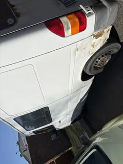 Biały furgon z widoczną rdzą na tylnej części, częściowo widoczny inny samochód po lewej stronie, ceglany budynek i trawa na tle, jasne niebo.