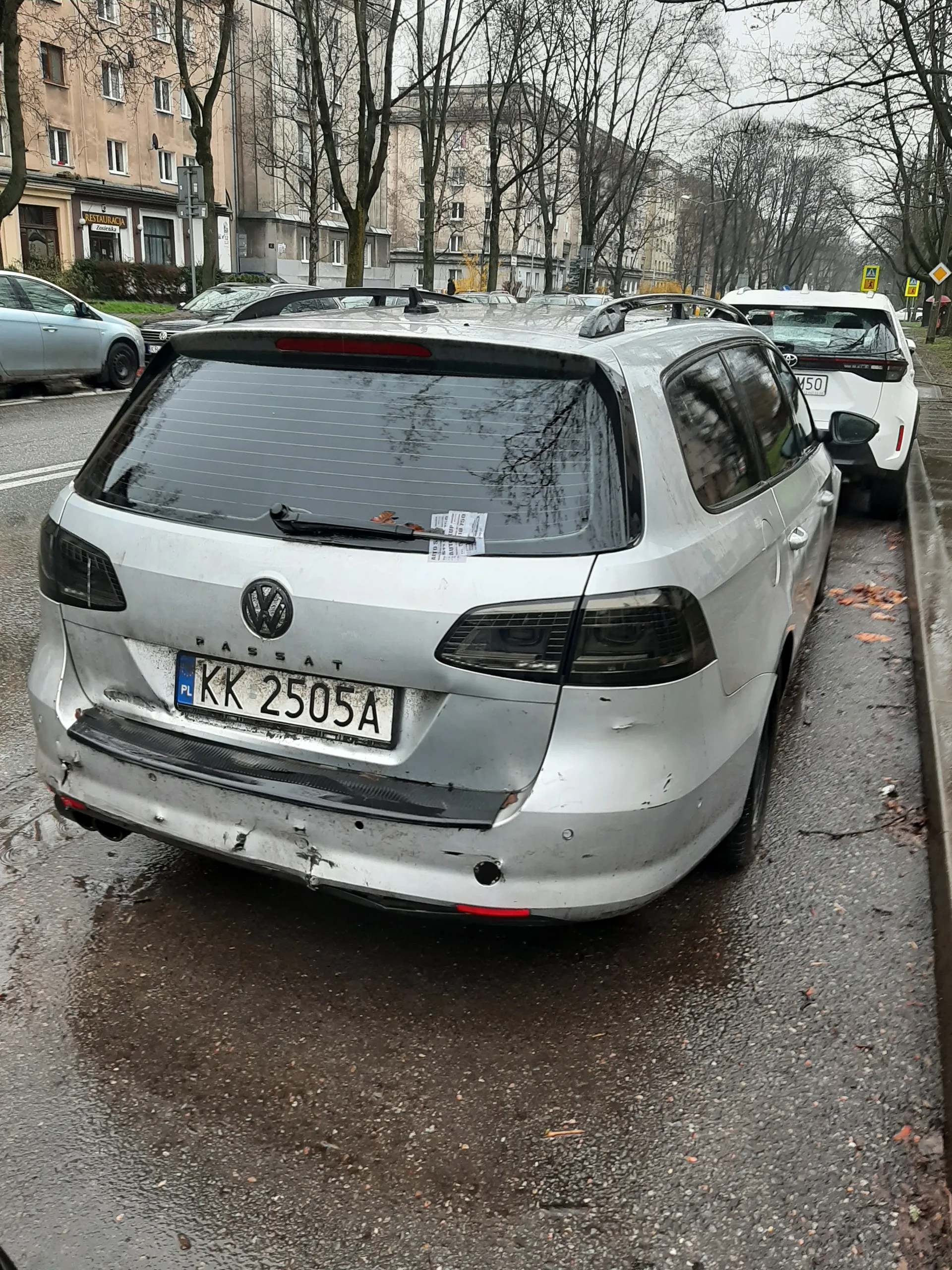 Samochód osobowy marki Volkswagen Passat w kolorze srebrnym z uszkodzonym tylnym zderzakiem, zaparkowany na mokrej nawierzchni drogi. Widoczna tablica rejestracyjna KK 2505A z niebieskim pasem po lewej stronie. Na tle budynki mieszkalne, drzewa bez liści, inne zaparkowane pojazdy oraz znaki drogowe. Na szybie tylnej naklejka. Nawierzchnia drogi mokra, widoczne opadłe liście. Niebo zachmurzone.
