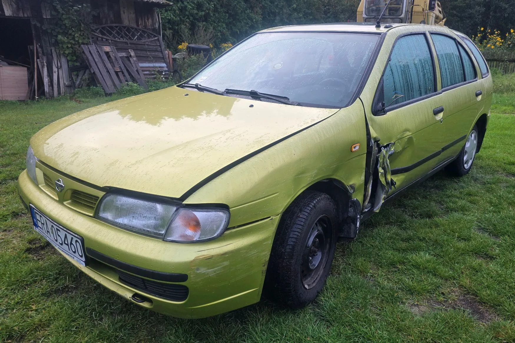 Jasnozielony samochód osobowy typu kombi zaparkowany na trawie. Pojazd posiada widoczne uszkodzenia karoserii po lewej stronie (prawa strona z perspektywy kierowcy), w tym wgniecenie drzwi kierowcy oraz uszkodzony błotnik. Na przednim zderzaku znajduje się tablica rejestracyjna o numerze ERA 05460. W tle widoczna jest drewniana szopa oraz zieleń.