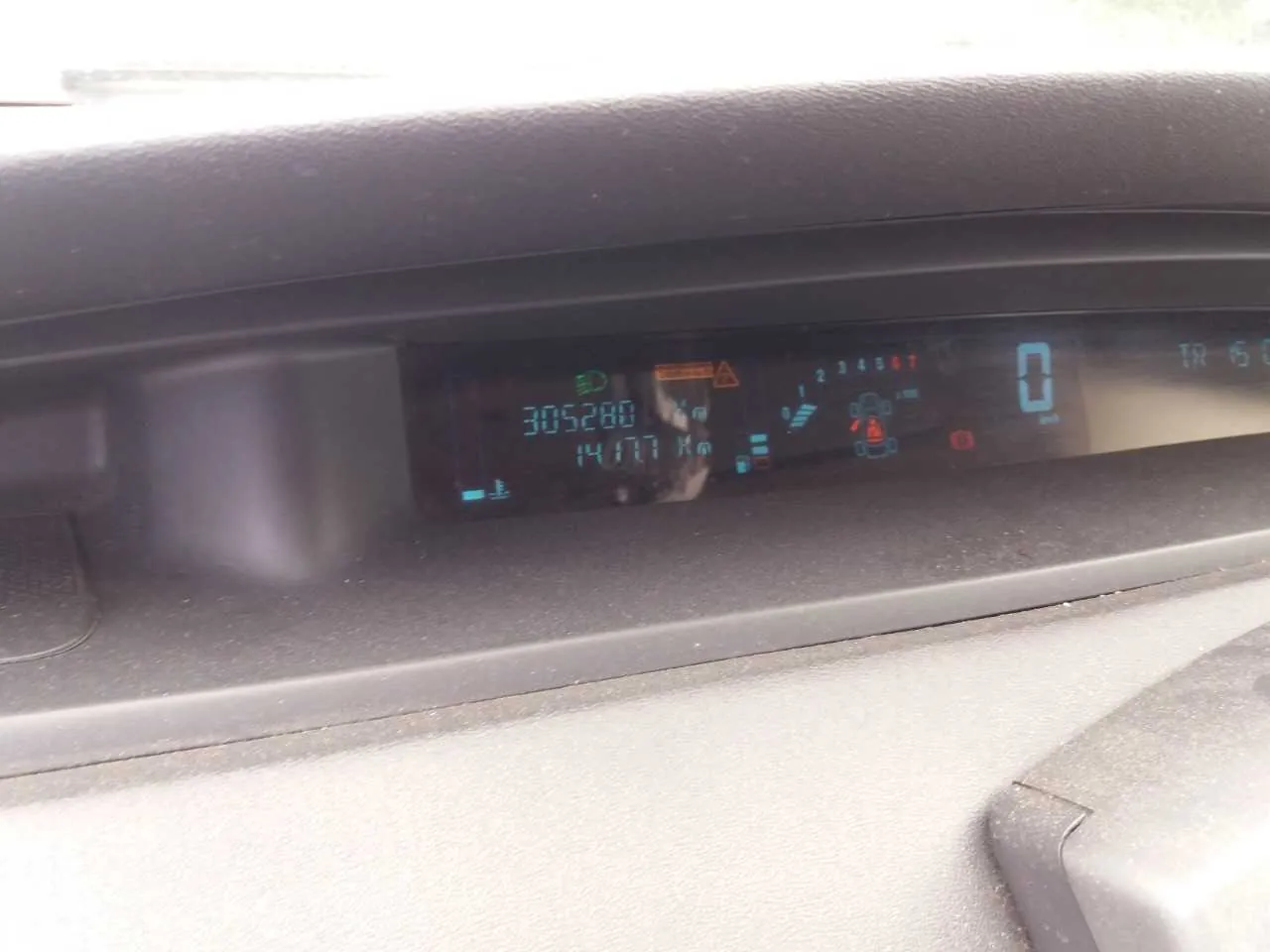 Zbliżenie na cyfrowy zegar samochodowy. Na wyświetlaczu widoczne są liczby: 305280 (przebieg), 14177 Km (przebieg podróży), 0 (prędkość). Obecne są również ikony: pas bezpieczeństwa, ostrzeżenie o lampie, wskaźnik biegu P, poziom paliwa i inne symbole ostrzegawcze. Wyświetlacz ma niebieskie i pomarańczowe oznaczenia na czarnym tle. W tle widoczna część deski rozdzielczej w kolorze szarym.