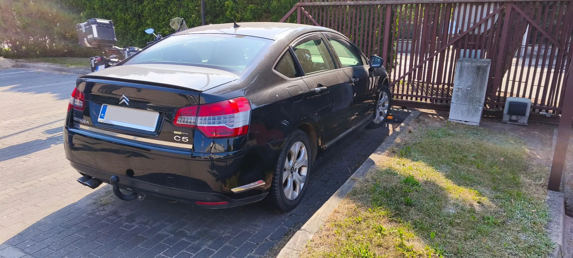 Czarny samochód marki Citroën C5 zaparkowany tyłem na kostce brukowej obok metalowej bramy. Na klapie bagażnika widoczne jest logo marki, napis C5 oraz tablica rejestracyjna z oznaczeniem PL. Samochód posiada zamontowany hak holowniczy.