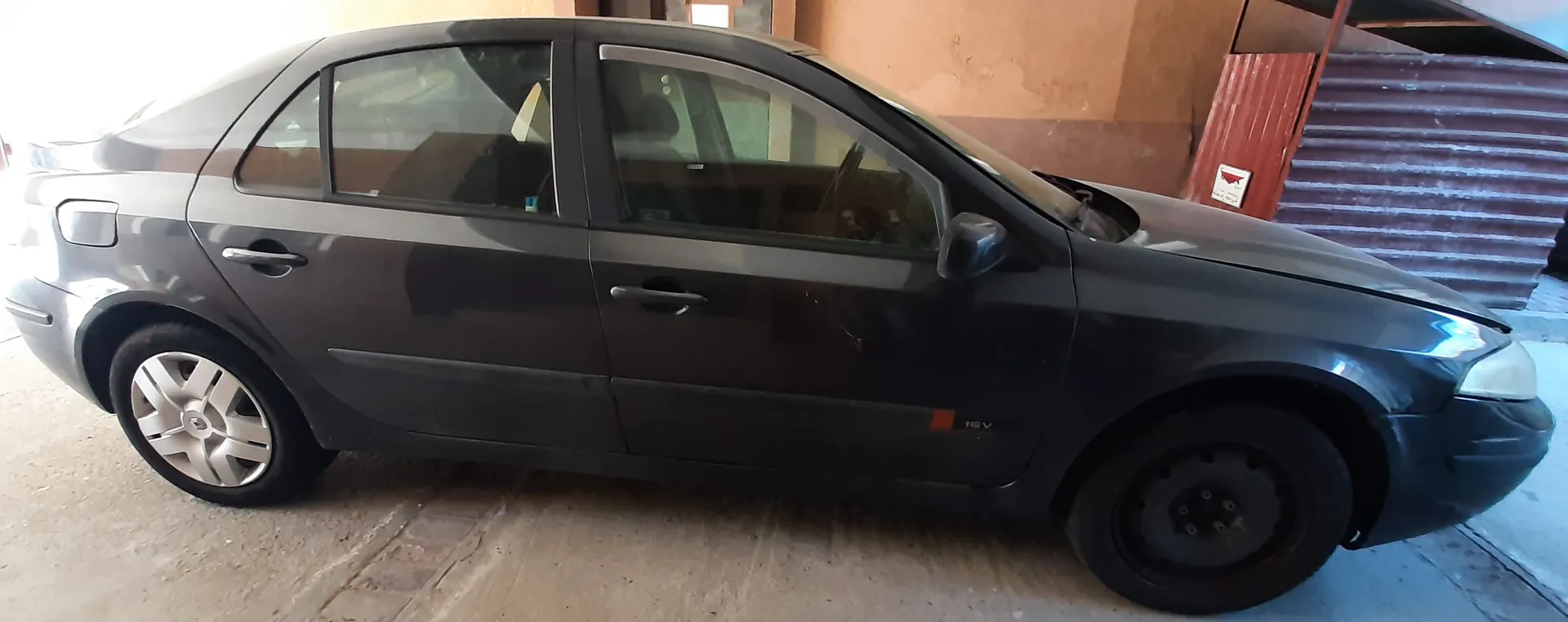 Ciemnoszary samochód osobowy typu sedan z czterema drzwiami zaparkowany na betonowej powierzchni. Widoczne są srebrne felgi na tylnym kole i czarne felgi stalowe na przednim kole. Na przednim błotniku znajduje się napis '16V'. W tle budynki z brązową ścianą i czerwoną metalową konstrukcją, a także tabliczka z czerwonym trójkątem.