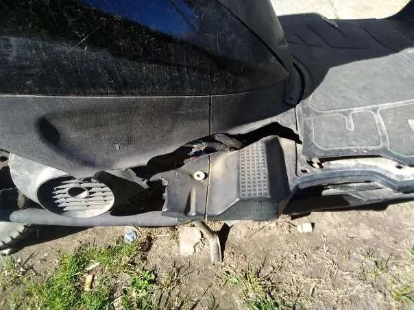 Zdjęcie przedstawia uszkodzoną część czarnego pojazdu, prawdopodobnie skutera lub motocykla. Widoczna jest pęknięta obudowa z bocznym wydechem o okrągłym kształcie z siatką, uszkodzony panel podłogowy z teksturą i śladami zniszczenia. Tło stanowi ziemia z trawą i drobnymi śmieciem. Brak czytelnego tekstu.