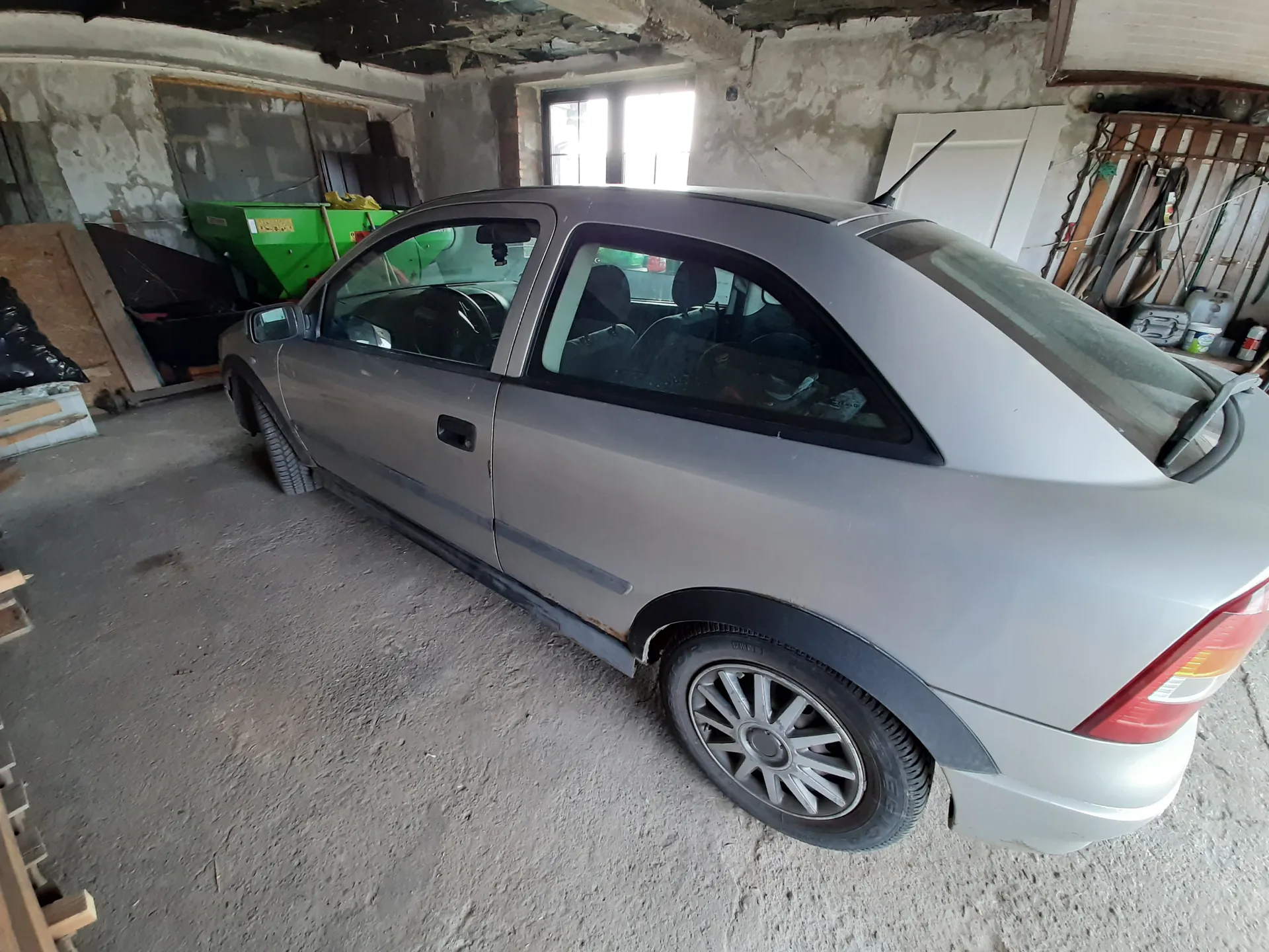 Srebrny samochód osobowy typu hatchback z dwoma drzwiami zaparkowany wewnątrz zaniedbanego garażu. Wnętrze garażu ma betonową podłogę, pęknięte i obłupione ściany oraz uszkodzone sufit. W tle widoczne są zielone pojemniki, okno wpuszczające światło oraz narzędzia wiszące na ścianie po prawej stronie. Samochód posiada czarne obwody okien, felgi ze srebrnymi kołpakami i czerwone światła tylnego. Wewnątrz pojazdu widoczne są siedzenia i przedmioty na tylnym siedzeniu.