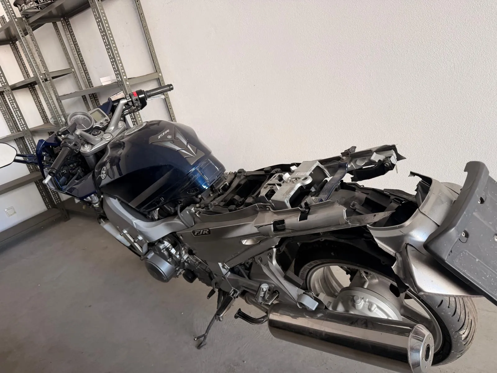 Motocykl marki Yamaha FJR w kolorze ciemnoniebieskim i srebrnym, pozbawiony tylnych owiewek i siedzenia, co odsłania ramę i elementy konstrukcyjne. Pojazd stoi na stopce w pomieszczeniu obok metalowego regału. Na baku widoczne jest logo FJR.