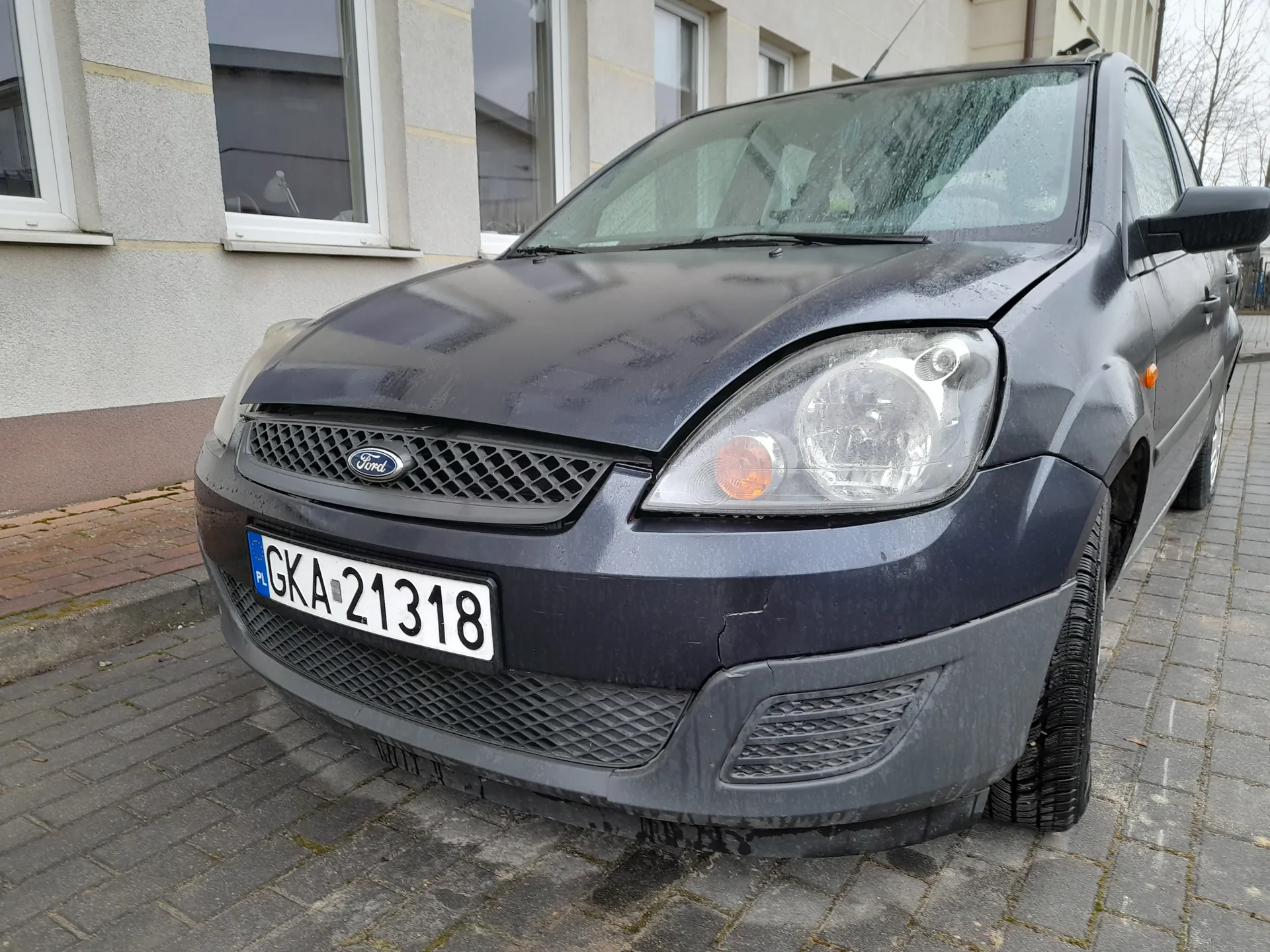 Ciemnoszary samochód marki Ford zaparkowany na brukowanej nawierzchni przed budynkiem z oknami. Widoczna tablica rejestracyjna z białym tłem i czarnym napisem GKA-21318 oraz niebieskim pasem z literami PL po lewej stronie. Na masce samochodu znajduje się logo Ford. Budynek w tle ma szare ściany i białe obramowania okien. Nawierzchnia składa się z szarych i czerwonych płytek brukowych.