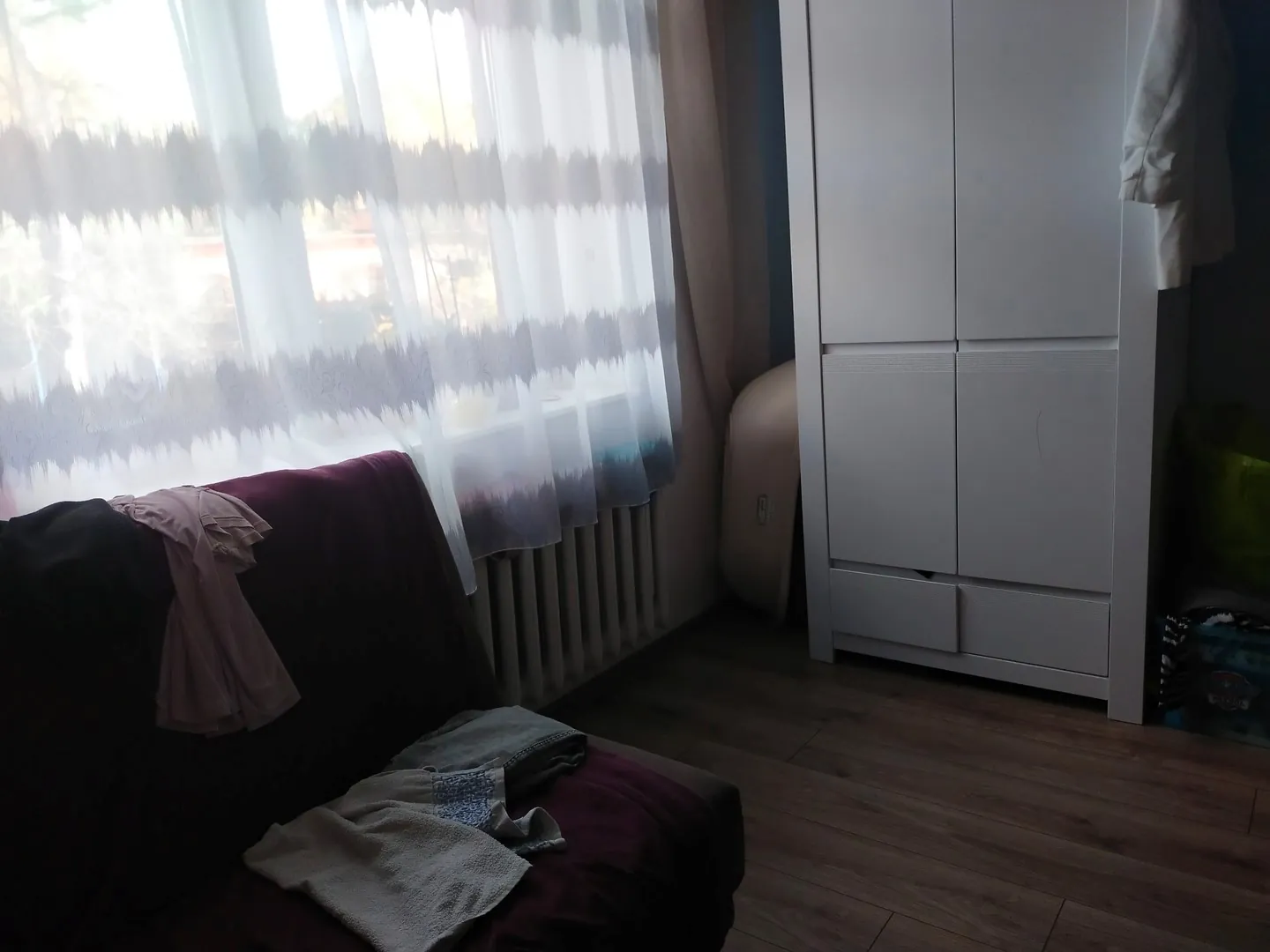 Wnętrze pokoju z ciemnofioletową kanapą w lewym dolnym rogu, na której leżą jasne ubrania. W tle znajduje się okno z białymi, półprzezroczystymi firankami, pod którym widoczny jest biały grzejnik. Po prawej stronie stoi wysoka, biała szafa dwudrzwiowa z szufladami na dole, a obok niej wisi biały szlafrok.