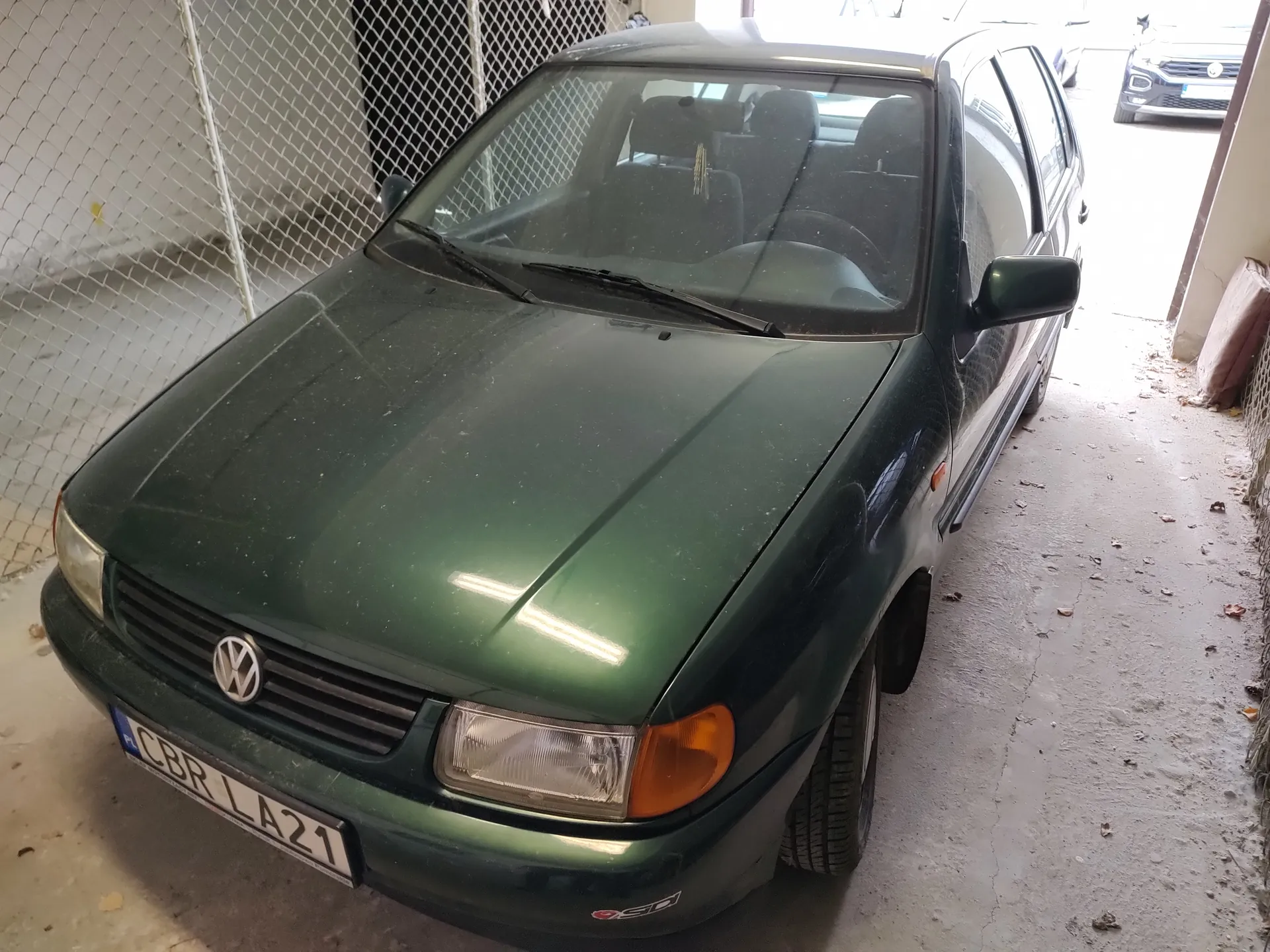 Zielony samochód marki Volkswagen zaparkowany w garażu. Widoczna tablica rejestracyjna CBR LA21, przednia szyba, reflektory oraz logo VW na masce. W tle siatka ogrodzeniowa, betonowa podłoga z rozrzuconymi liśćmi oraz częściowo widoczny inny samochód na zewnątrz garażu.